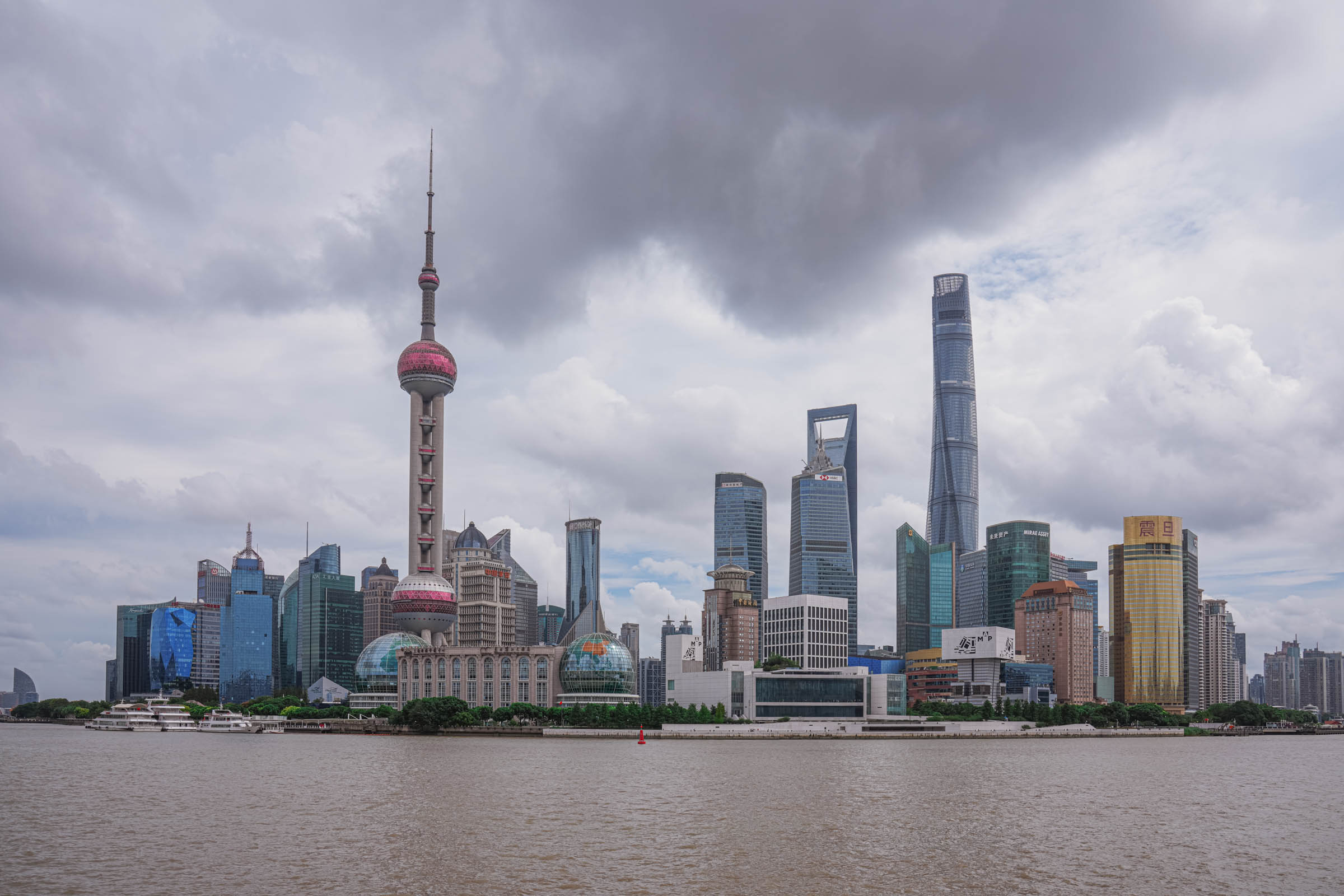 Breathtaking panorama of Shanghai from the Bund, featuring the distinctive Oriental Pearl Tower, the soaring Shanghai Tower, and the lofty Shanghai World Financial Center piercing a cloudy sky across the river.