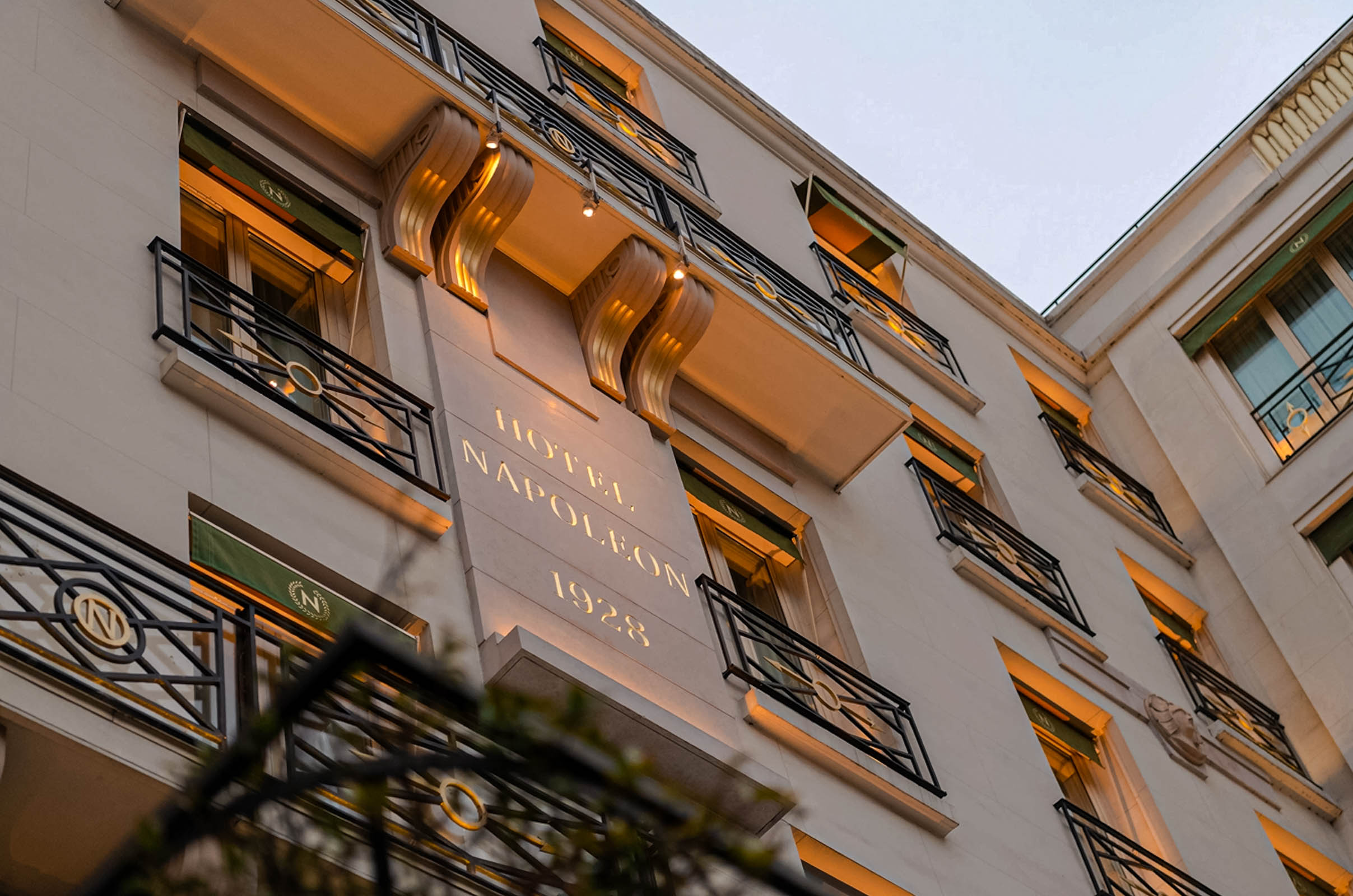 Illuminated facade of Hôtel Napoléon reading "HOTEL NAPOLEON 1928" at dusk, with "N" monograms on the wrought-iron balconies
