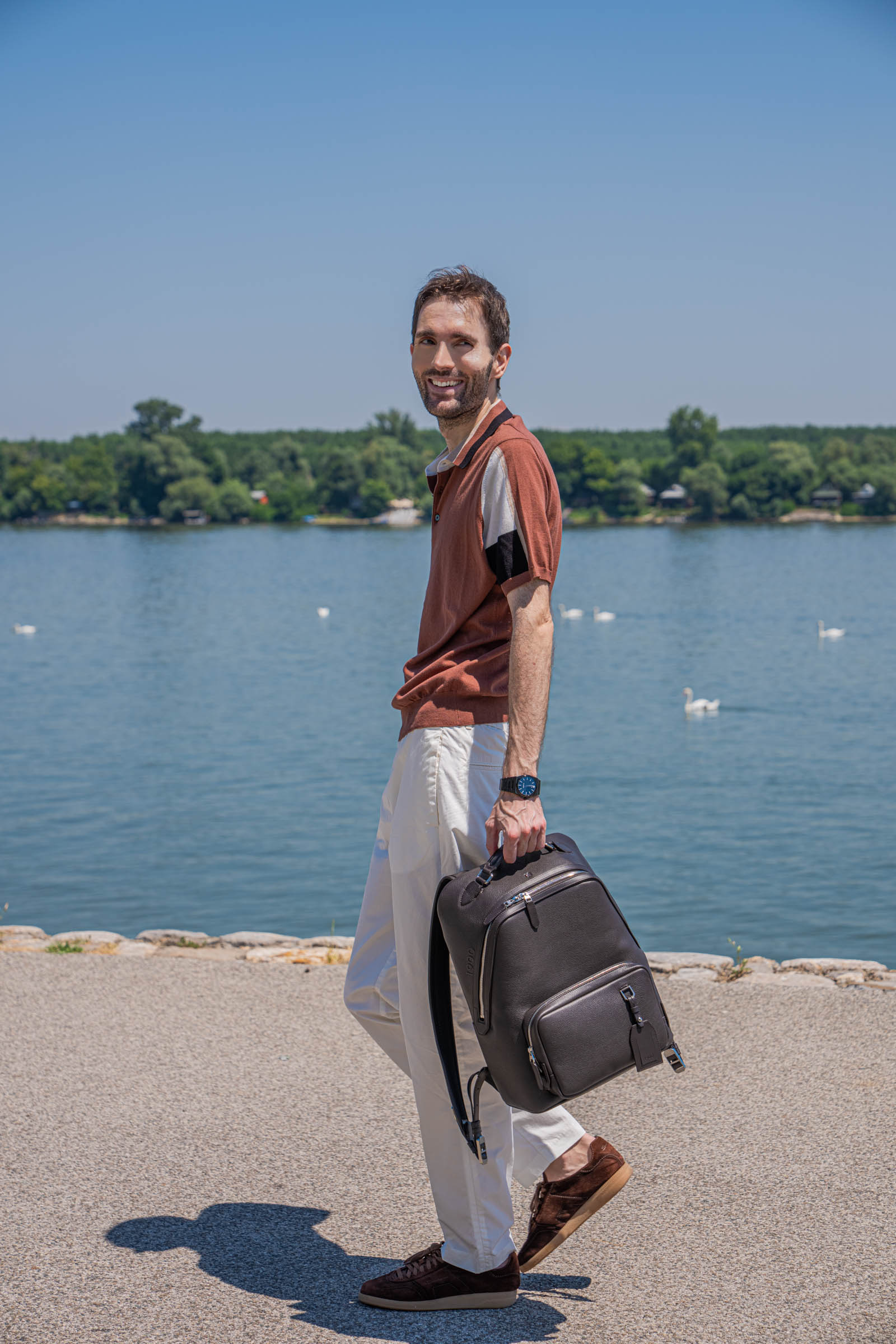 Marko Tadić, founder of Mr.M by Marko Tadić, walking along the Danube riverside promenade in Belgrade while glancing back over his shoulder and smiling, carrying the 1900 Tempo Grande dark brown leather backpack by its top handle, wearing a rust, cream and black Kiton knitted silk polo shirt with white Kiton cotton trousers and brown suede Santoni Oly sneakers, swans floating on the calm river behind him.