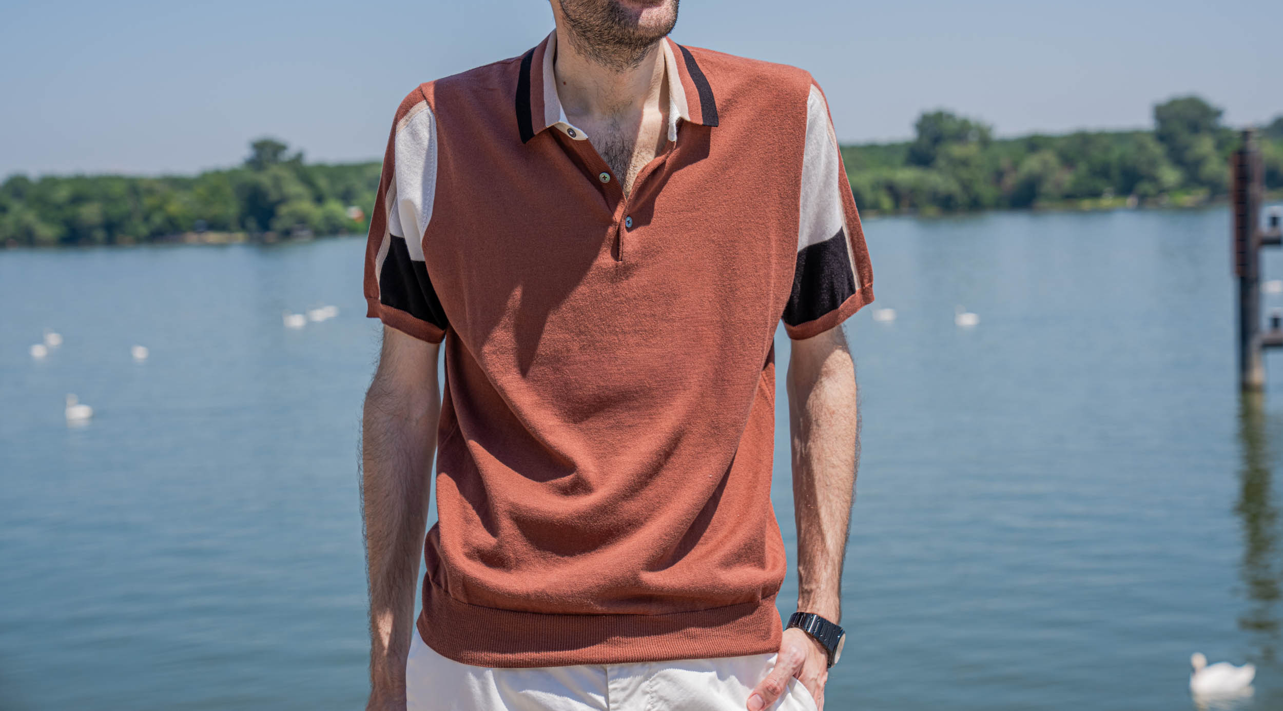 Close-up of the Kiton knitted silk polo shirt's rust, cream and black colorblock pattern and contrast-trimmed collar on Marko Tadić, the Danube river softly blurred behind.