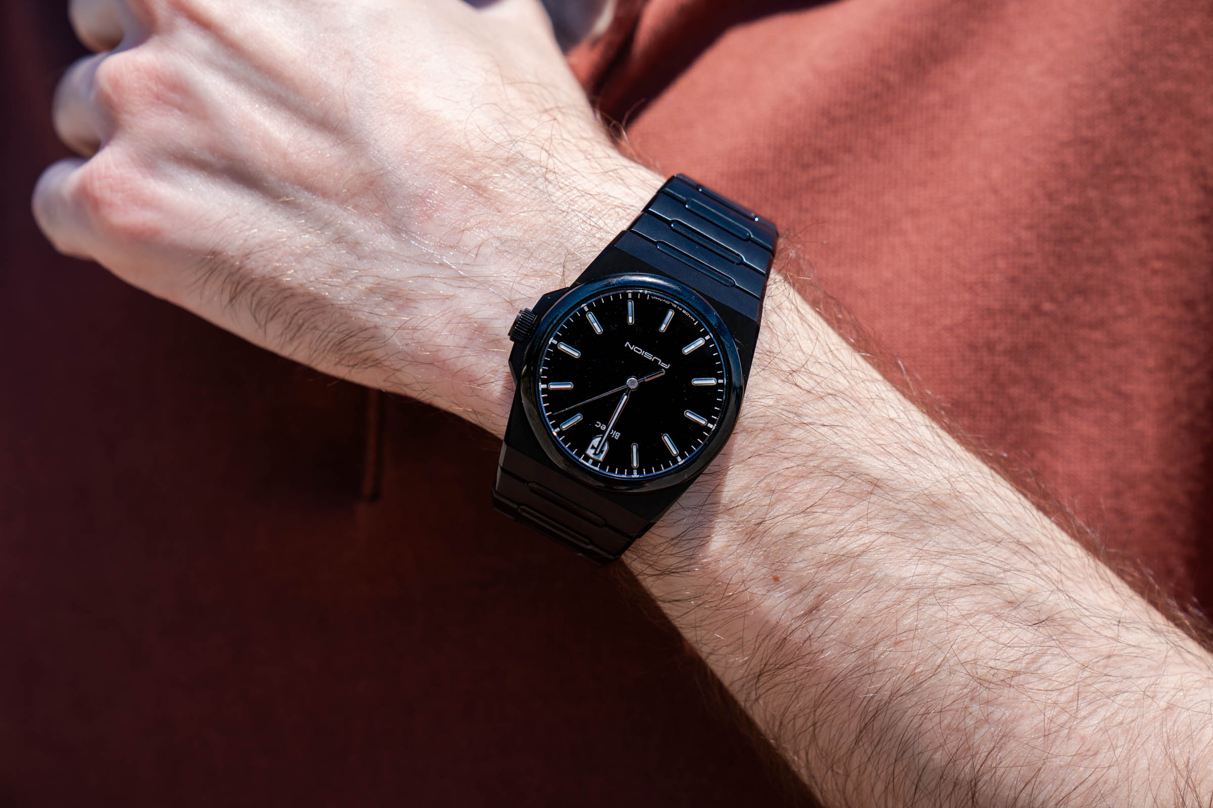 Extreme macro shot of the Biatec Fusion 03 watch's FUSION-branded black dial on Marko Tadić's wrist, set against the warm rust-toned knit texture of his Kiton silk polo.