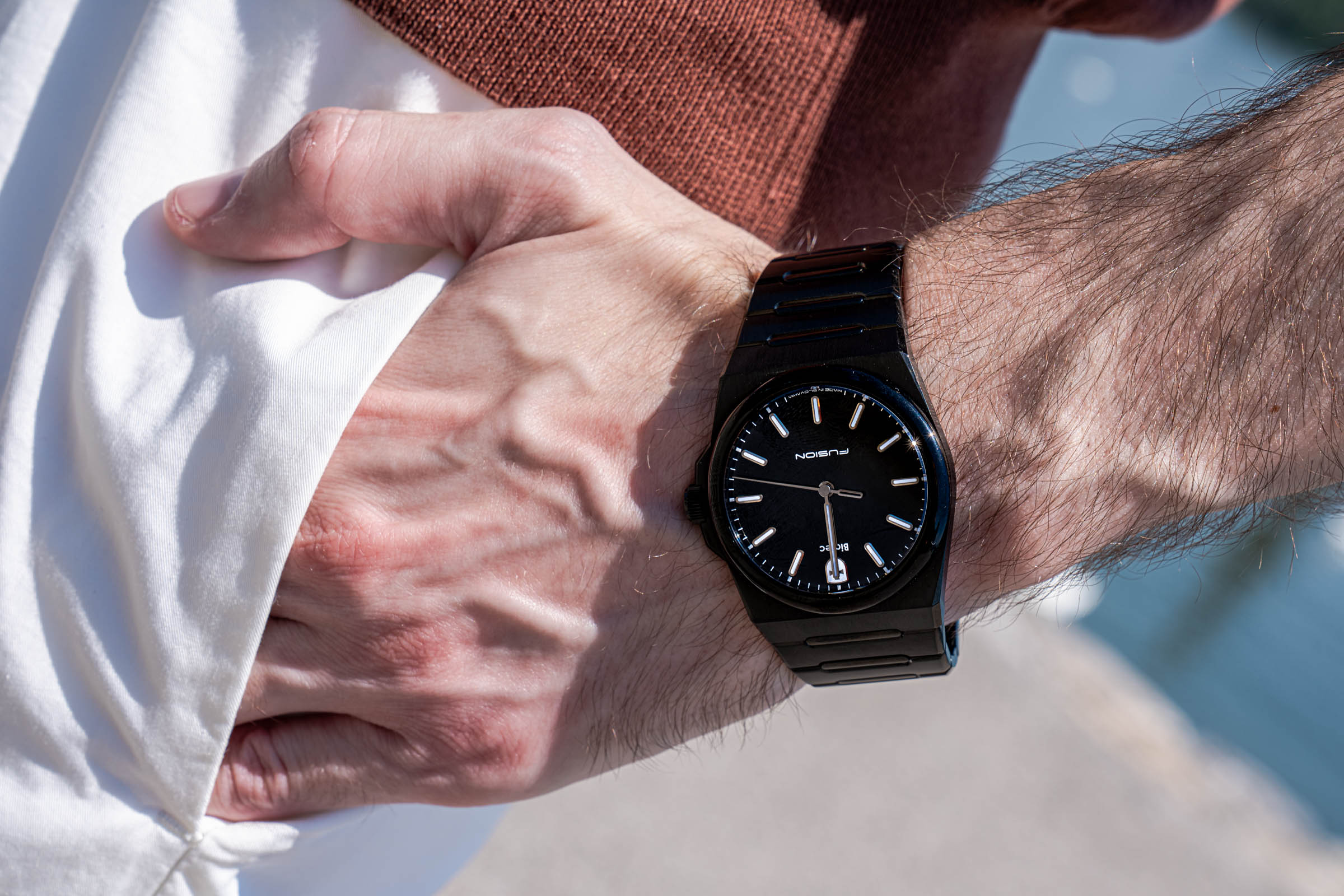 Detailed macro close-up of the Biatec Fusion 03 watch on Marko Tadić's wrist as he gathers his white Kiton trouser fabric in the same hand, soft blue Danube water blurred in the corner of the frame.