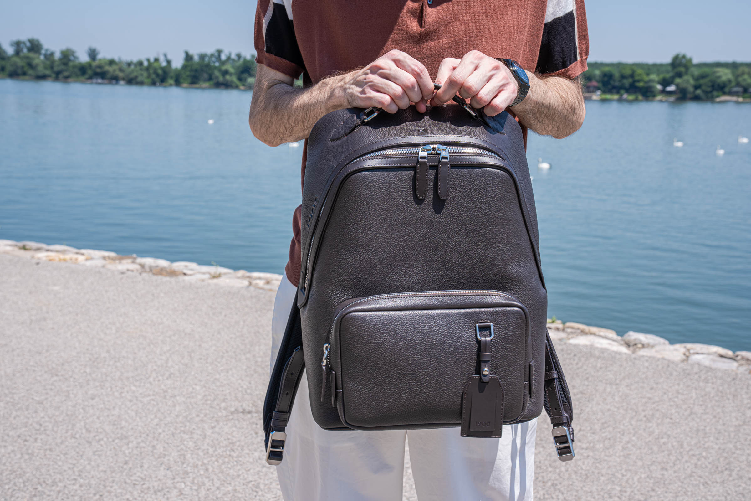 Close-up of Marko Tadić's hands gripping the top handle of the 1900 Tempo Grande backpack, the leather 1900 luggage tag and front zip pocket in sharp focus, his patterned Kiton polo visible above.