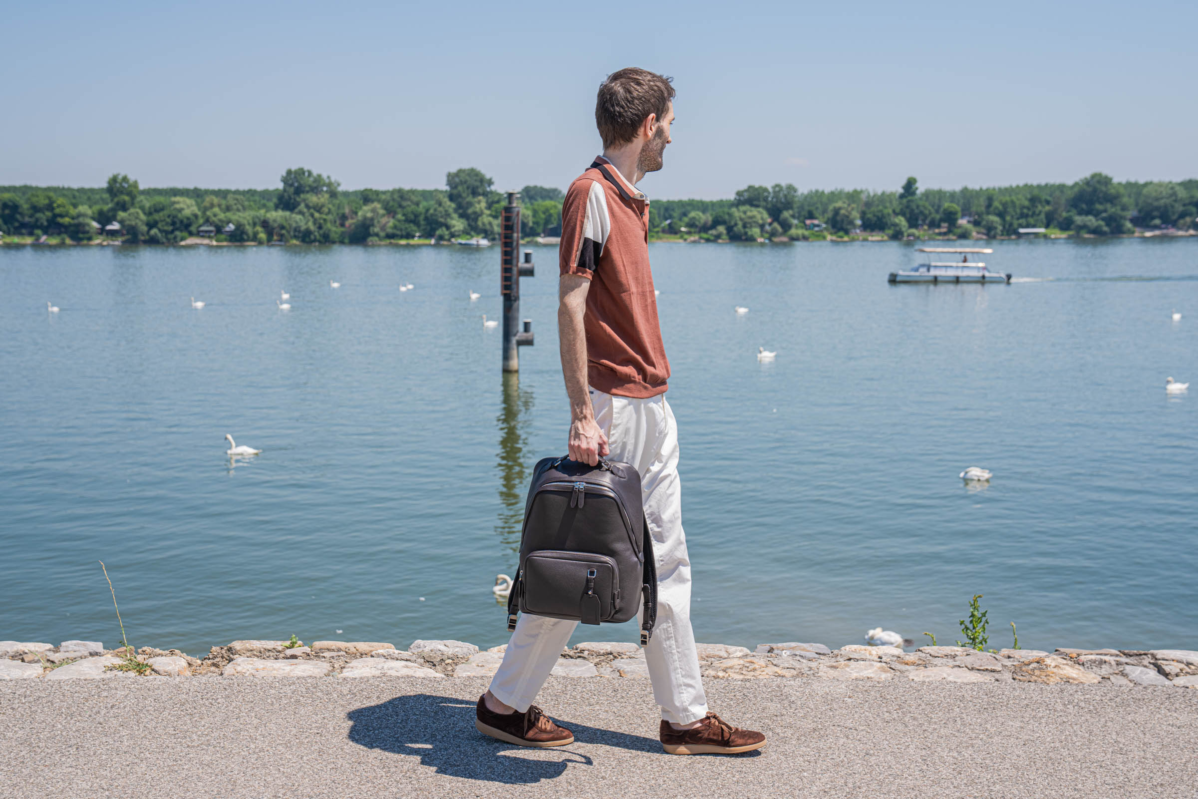 Marko Tadić walking away in profile past an old wooden dock winch and moored boats, holding the 1900 Tempo Grande leather backpack, a lone swan swimming nearby, his Kiton polo and white trousers finished with brown suede Santoni Oly sneakers.