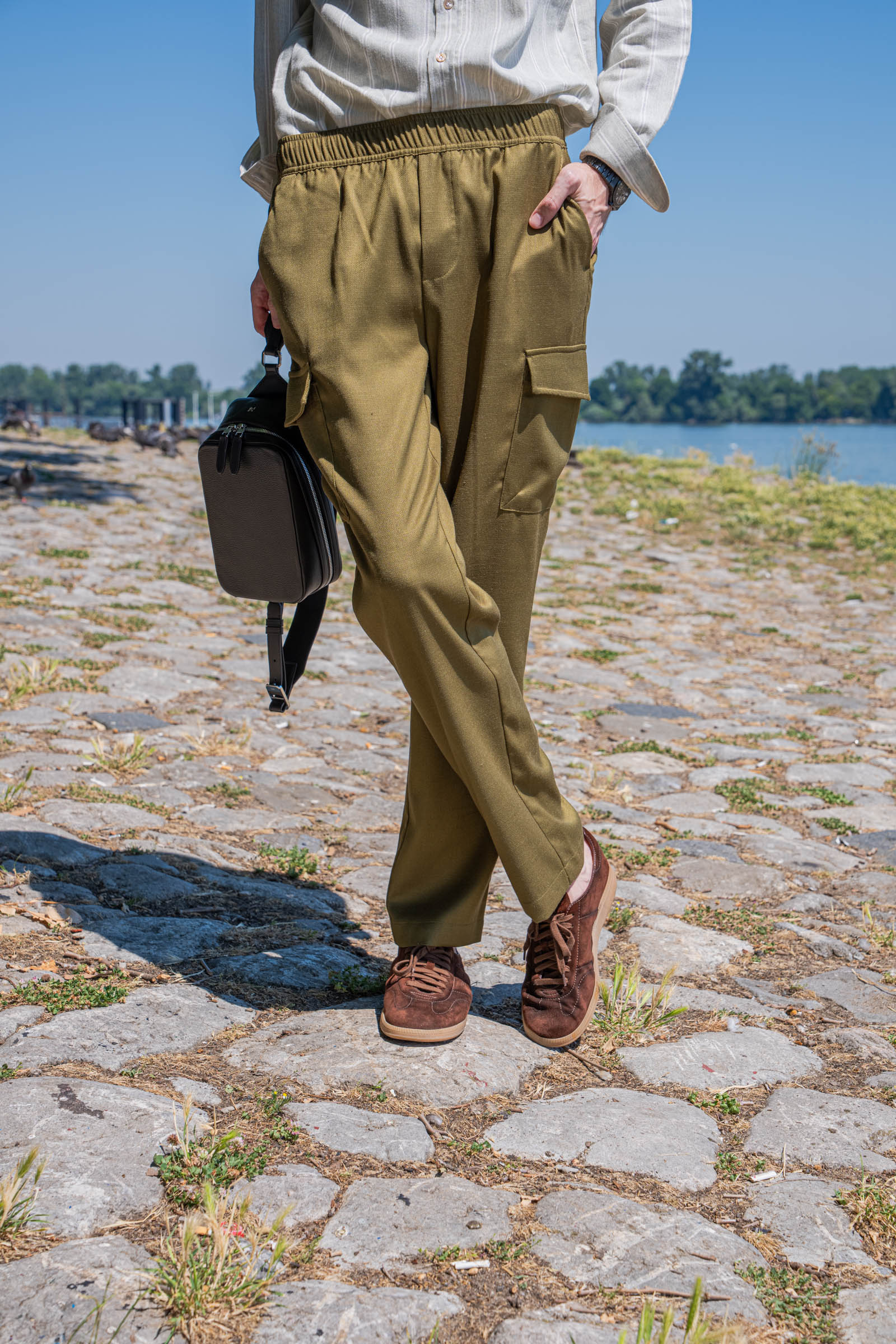 Lower body detail of men's summer style: Loro Piana olive linen-silk cargo trousers with side flap pocket, Santoni Oly dark brown suede sneakers and the 1900 grained leather crossbody bag held in hand on Zemun quay, Belgrade.