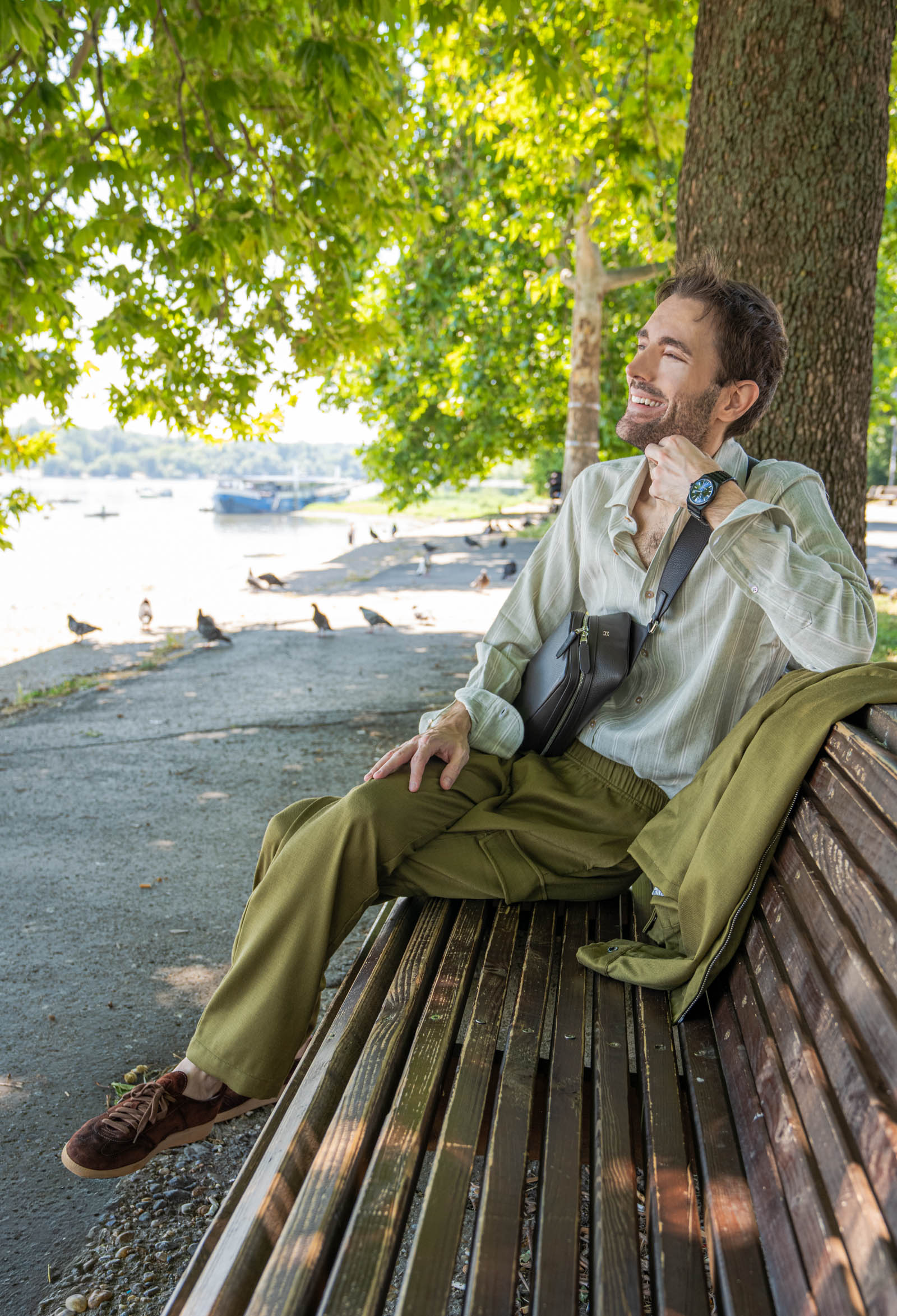Menswear blogger Marko Tadić rests on a park bench in Zemun wearing the full olive Loro Piana linen-silk set, with the 1900 grained leather crossbody bag placed beside him on the weathered wood.
