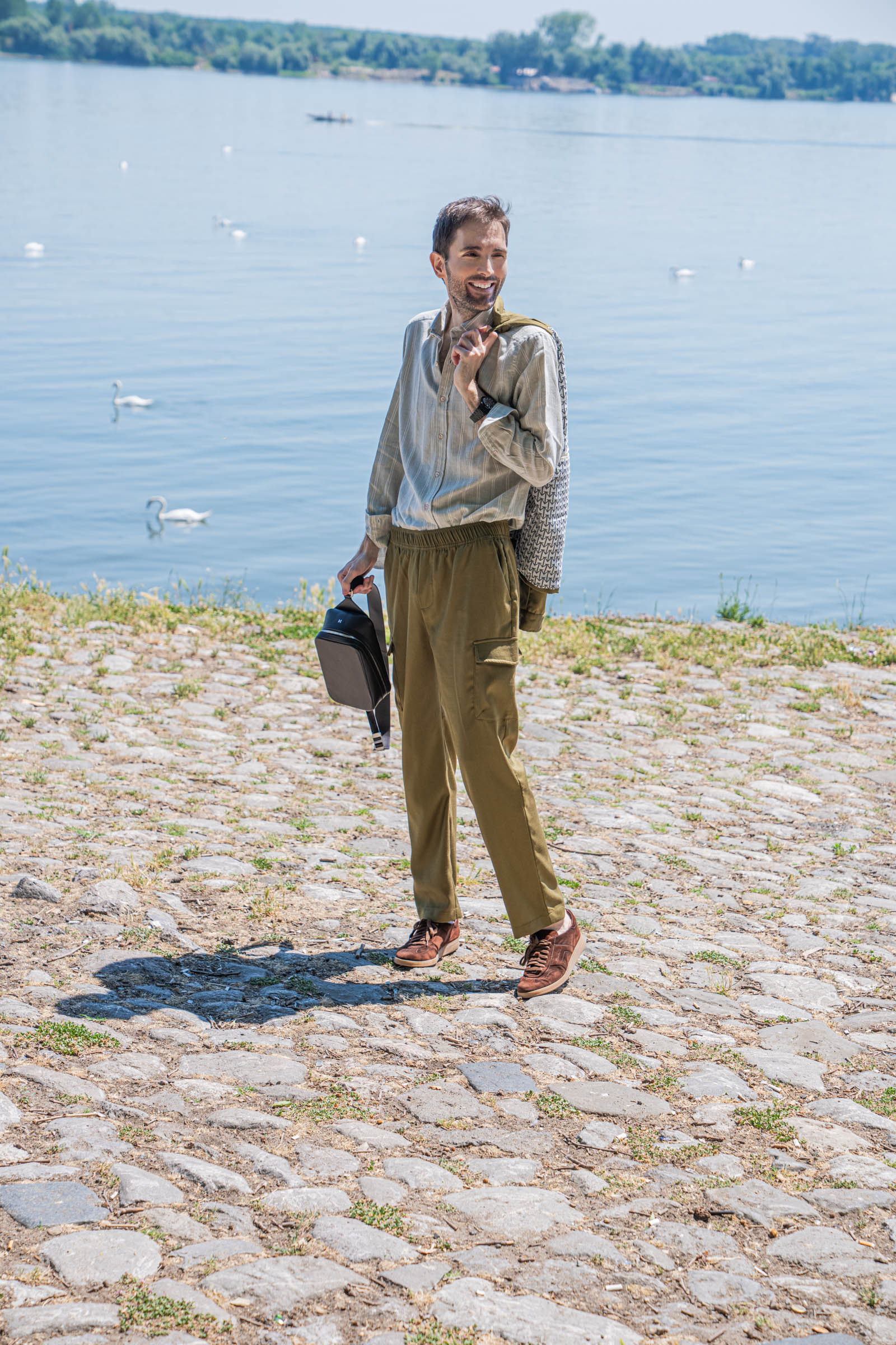 Side profile of fashion blogger Marko Tadić walking Zemun quay with a Loro Piana linen-silk jacket slung over his shoulder and the 1900 Tempo Grande crossbody bag in dark brown grained leather in his right hand.
