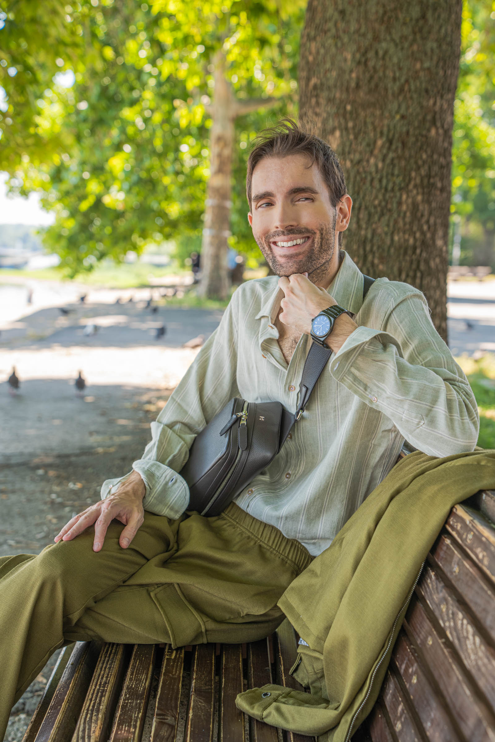 Smiling portrait of menswear blogger Marko Tadić in an olive green Loro Piana jacket with the 1900 Tempo Grande crossbody bag on his chest, the Danube river stretching behind him in Belgrade, Serbia.