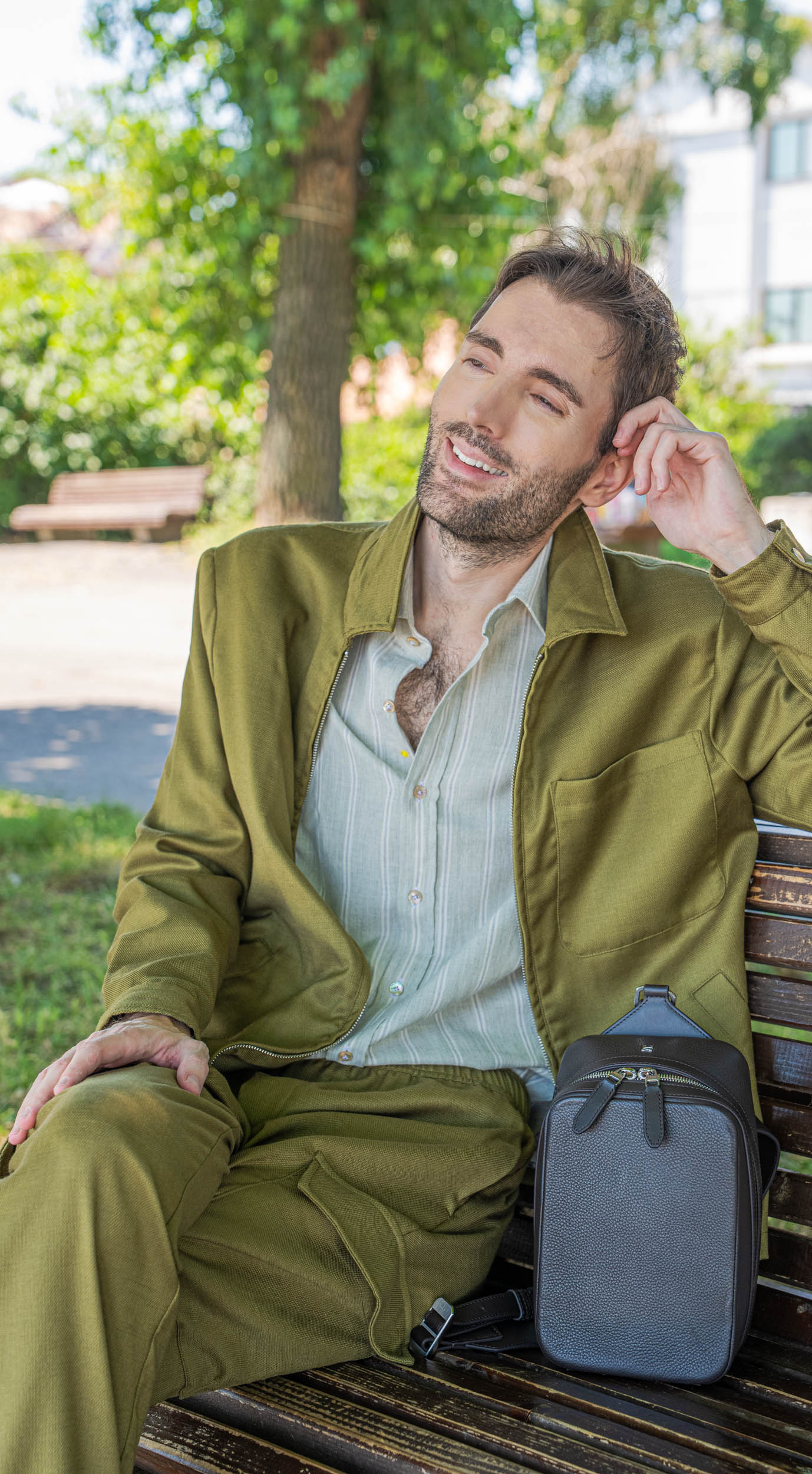 Menswear blogger Marko Tadić relaxes on a wooden bench under plane trees on Zemun quay, wearing the 1900 Tempo Grande crossbody bag across his chest with a striped Giannetto Portofino shirt and black Biatec watch.