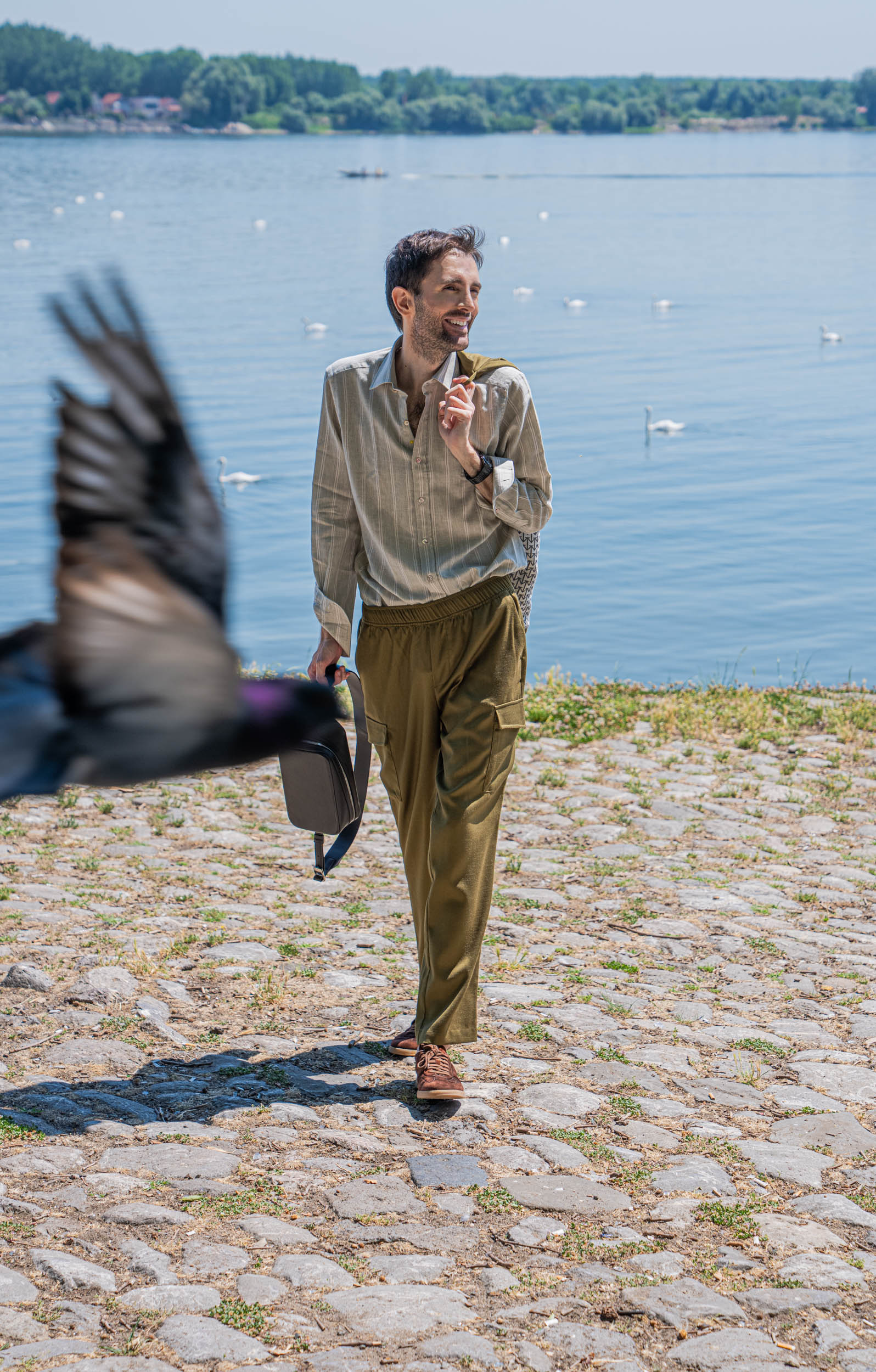A pigeon flies through the frame as Marko Tadić of Mr.M by Marko Tadic strolls along Zemun quay carrying the 1900 Tempo Grande crossbody bag, dressed in a striped Giannetto Portofino shirt and olive Loro Piana cargo trousers.