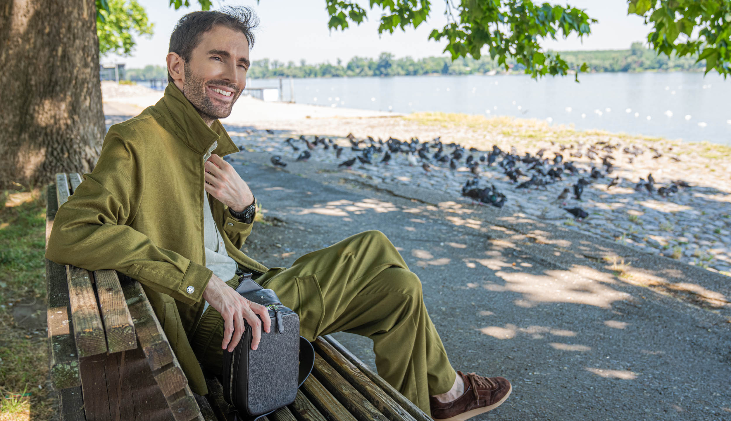 Marko Tadić of Mr.M by Marko Tadic sits on a riverside bench wearing the 1900 grained leather crossbody bag and a black Biatec Fusion 03 automatic watch, with his olive linen-silk jacket draped over the backrest.