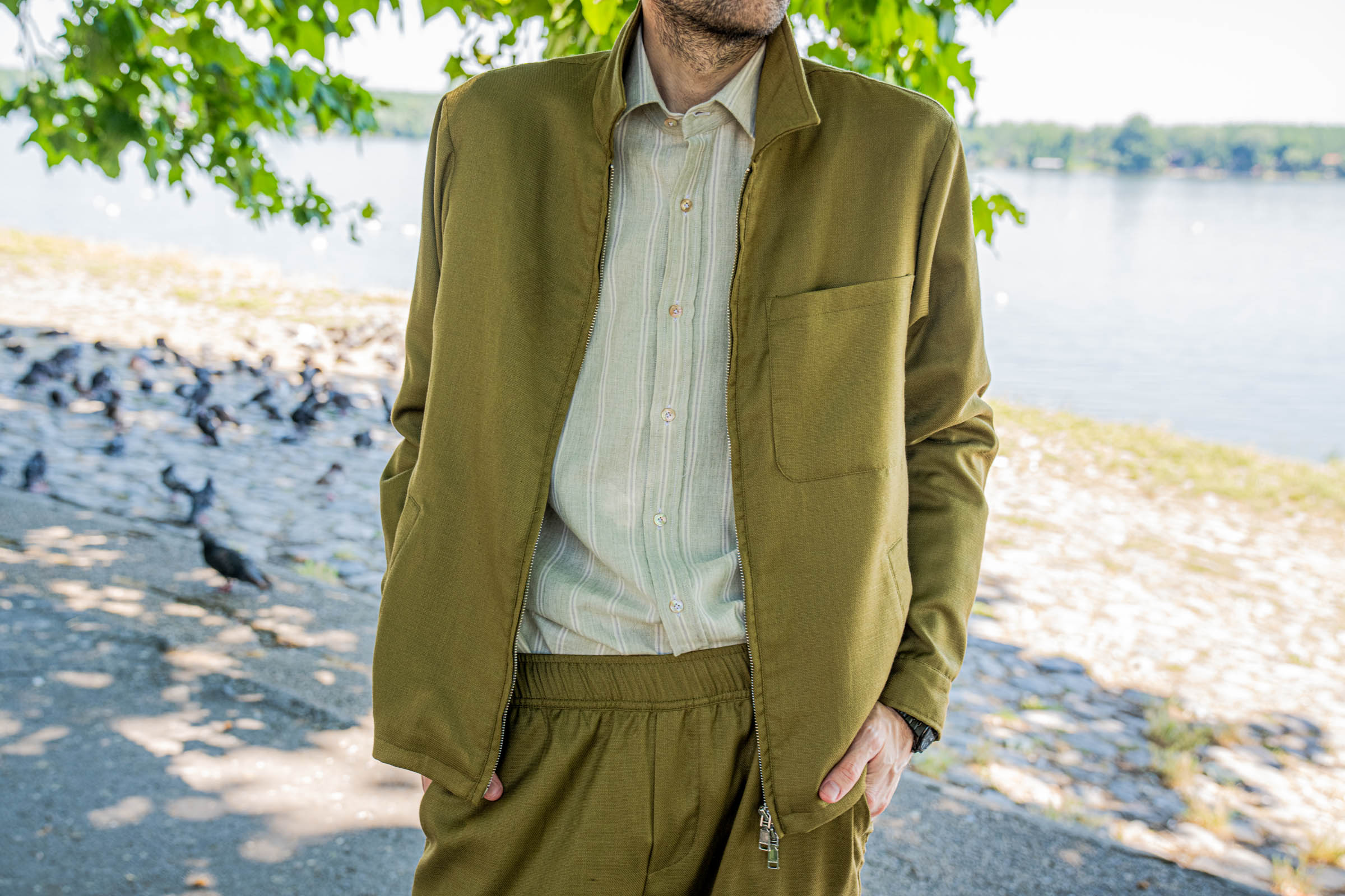 Torso detail of the Loro Piana olive linen-silk jacket worn half-zipped over a sage striped Giannetto Portofino shirt, photographed by the Danube river for the Mr.M by Marko Tadic quiet luxury editorial.