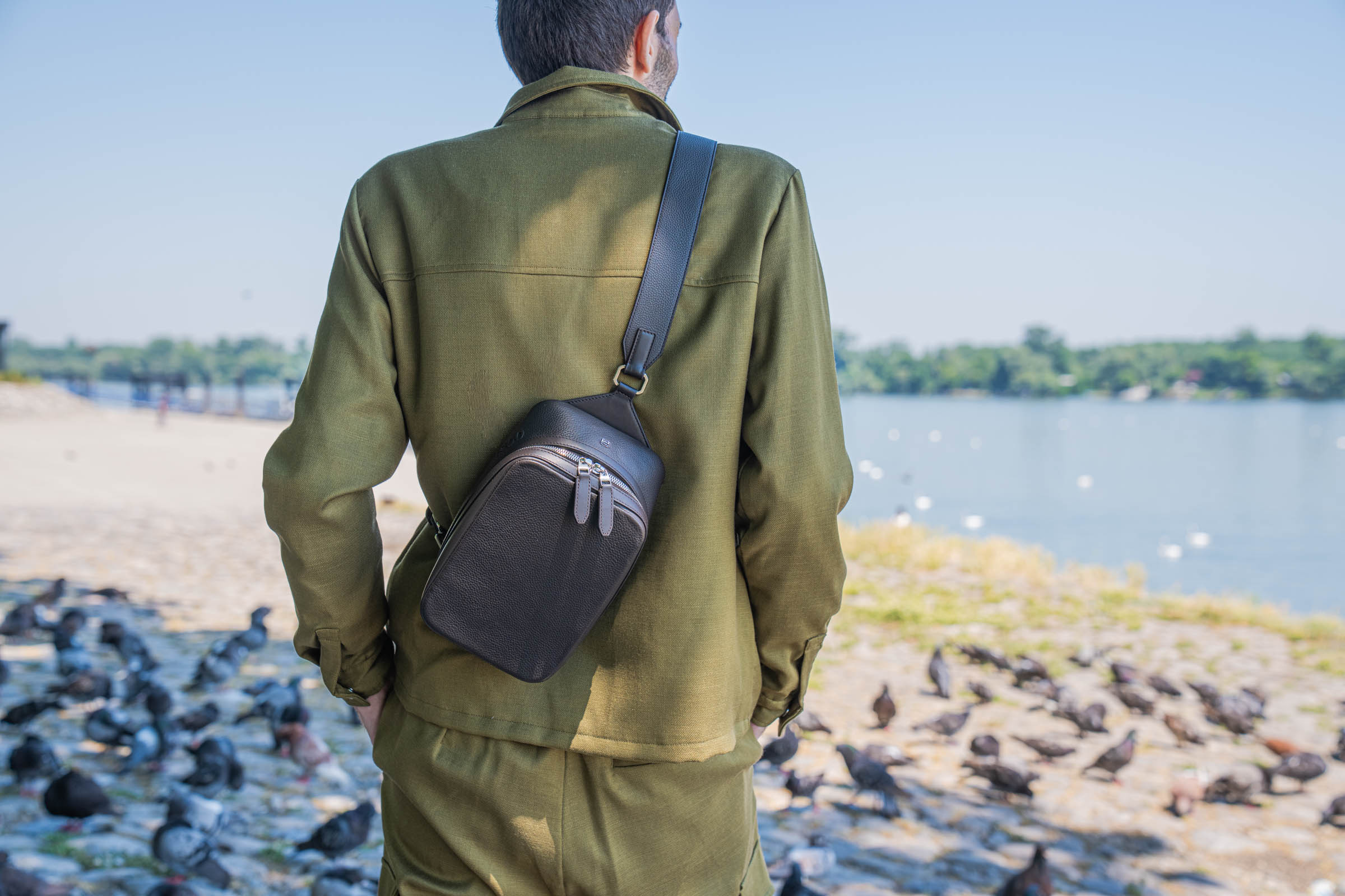 Back view of Marko Tadić wearing the 1900 Tempo Grande crossbody bag slung across his back over an olive Loro Piana linen-silk jacket, watching swans and pigeons along the Danube river at Zemun quay.