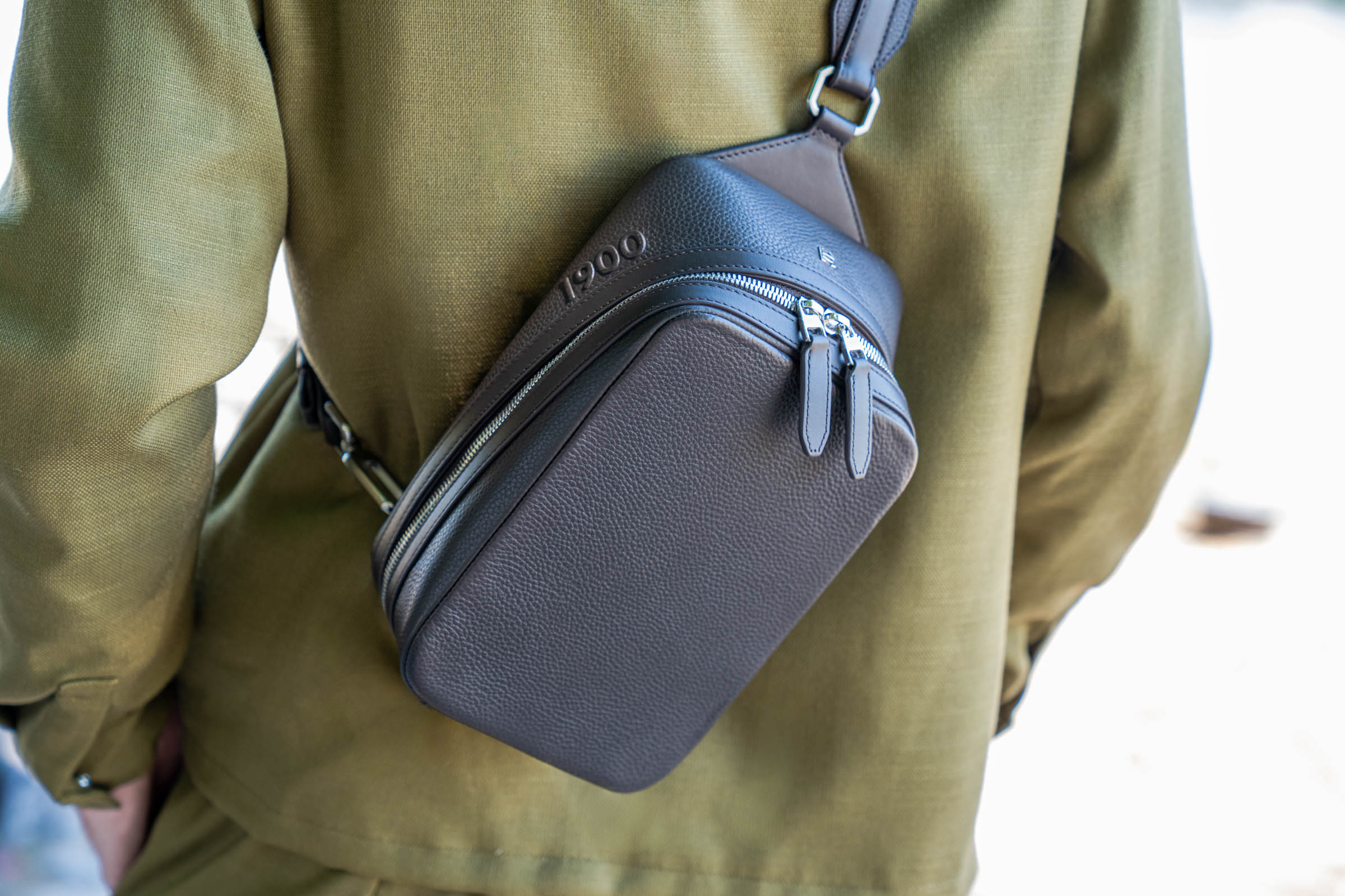 Detail of the 1900 Tempo Grande crossbody bag worn on the back, with the embossed 1900 lettering, brushed leather panel and silver hardware standing out against Marko Tadić's olive green jacket.