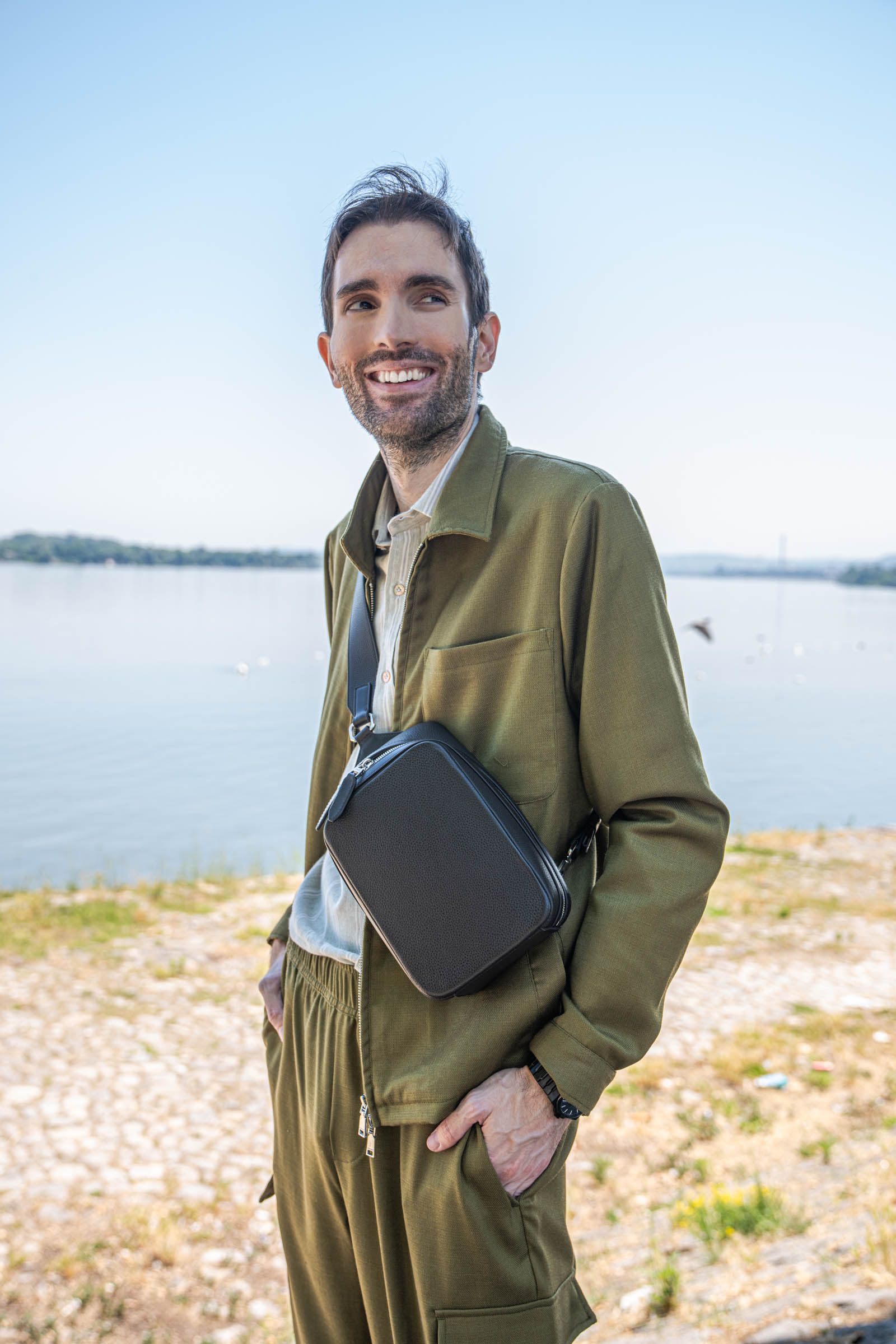 Marko Tadić, founder of Mr.M by Marko Tadic luxury blog, glances back smiling while carrying the 1900 crossbody bag along the Danube, wearing an olive Loro Piana set and Santoni Oly suede sneakers.