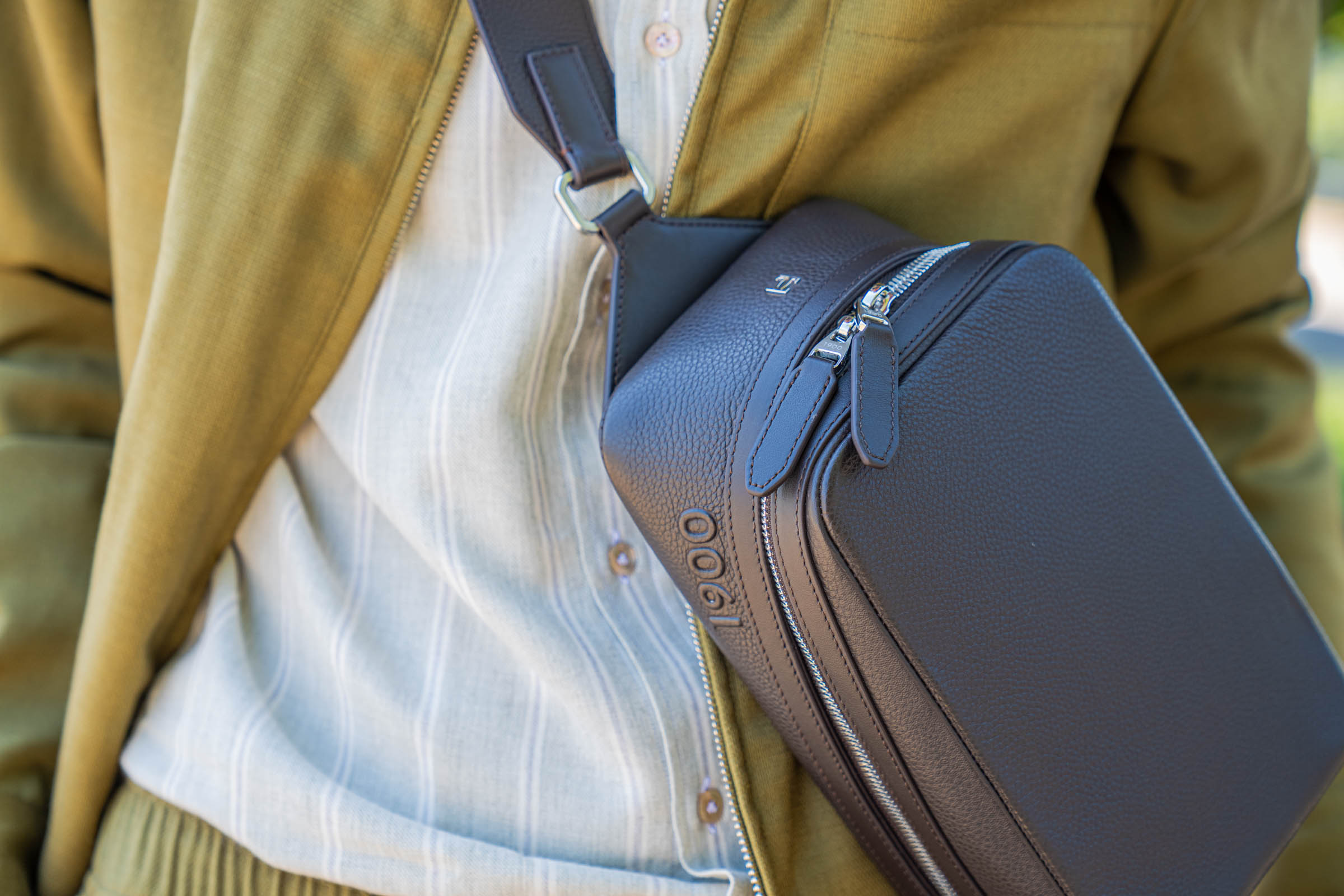 Detail of the dark brown 1900 Tempo Grande crossbody bag resting against olive green Loro Piana linen-silk cargo trousers, showing how earth tones and grained leather anchor a refined men's summer outfit.