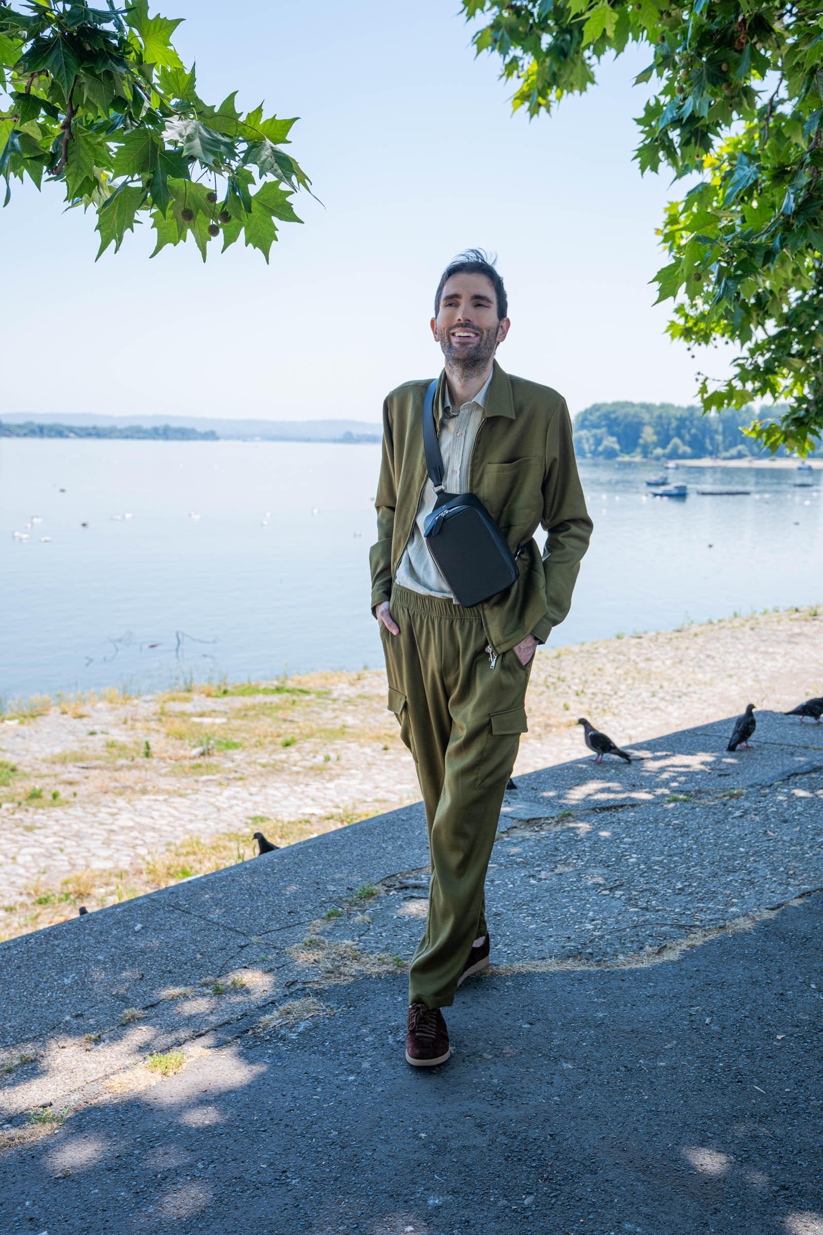 Marko Tadić, founder of the luxury menswear blog Mr.M by Marko Tadic, carries the 1900 Tempo Grande crossbody bag in dark brown grained leather while walking the cobblestone Zemun quay in Belgrade, with swans gliding on the Danube river behind him.