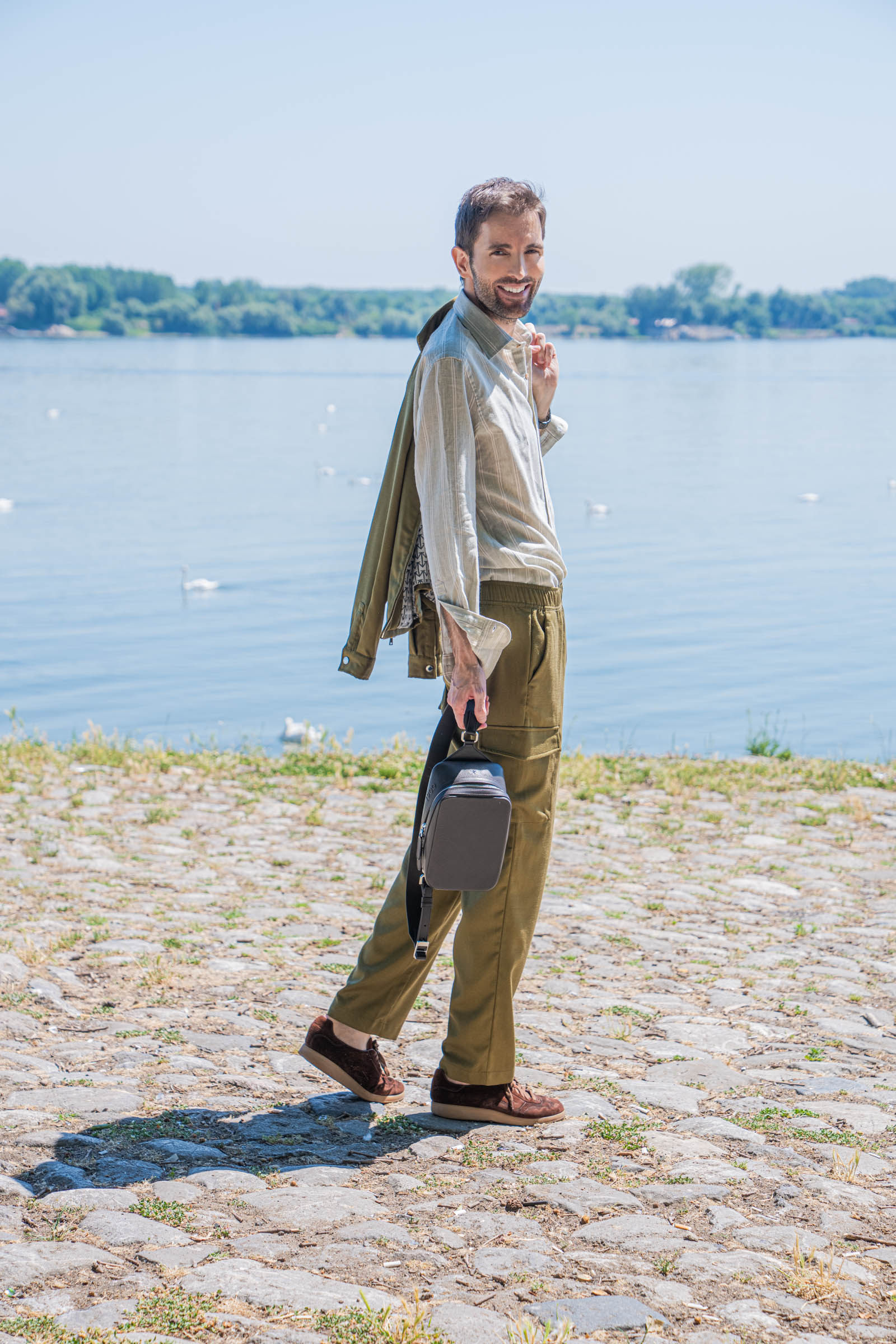 Marko Tadić, founder of Mr.M by Marko Tadic, stands on the sunlit Zemun quay holding the 1900 crossbody bag by its detachable strap, pairing a sage striped shirt with olive green linen-silk cargo trousers by Loro Piana.