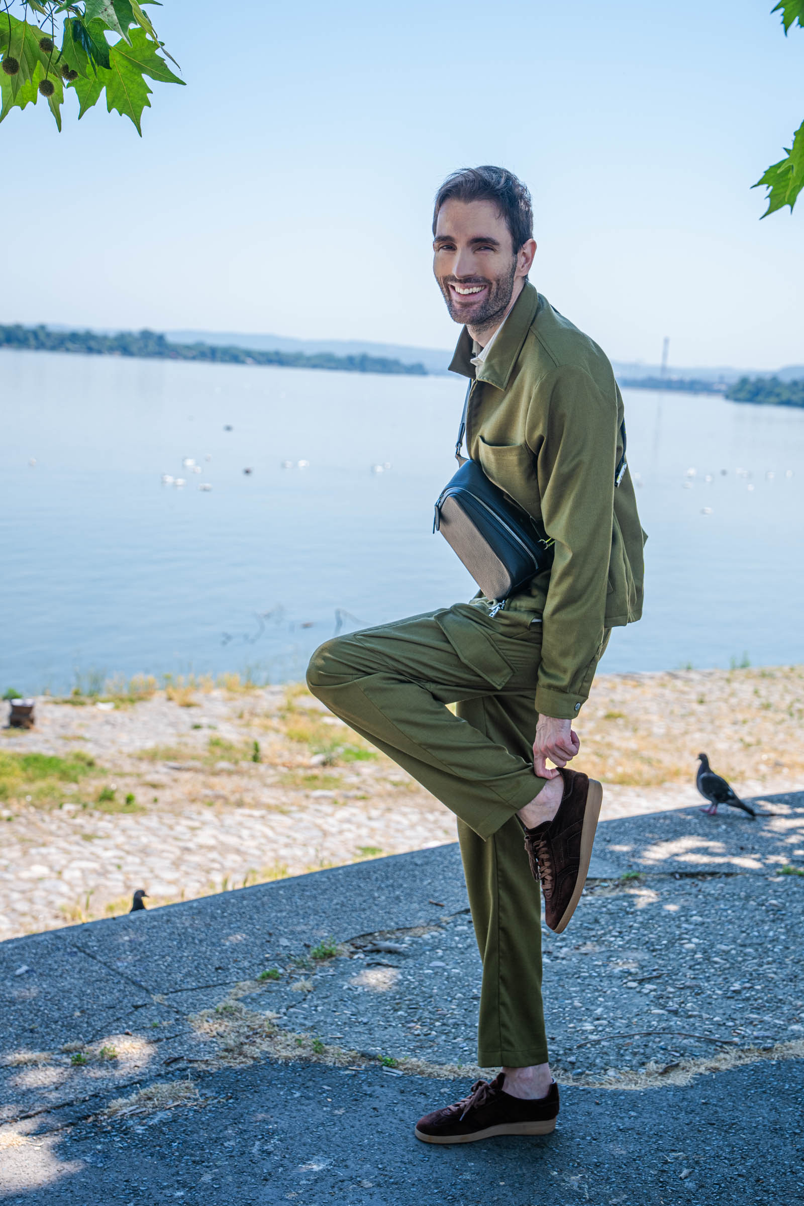 Marko Tadić, founder of Mr.M by Marko Tadic, walks under plane trees on Zemun quay in a full olive Loro Piana linen-silk set with the 1900 dark brown grained leather crossbody bag worn across his chest.