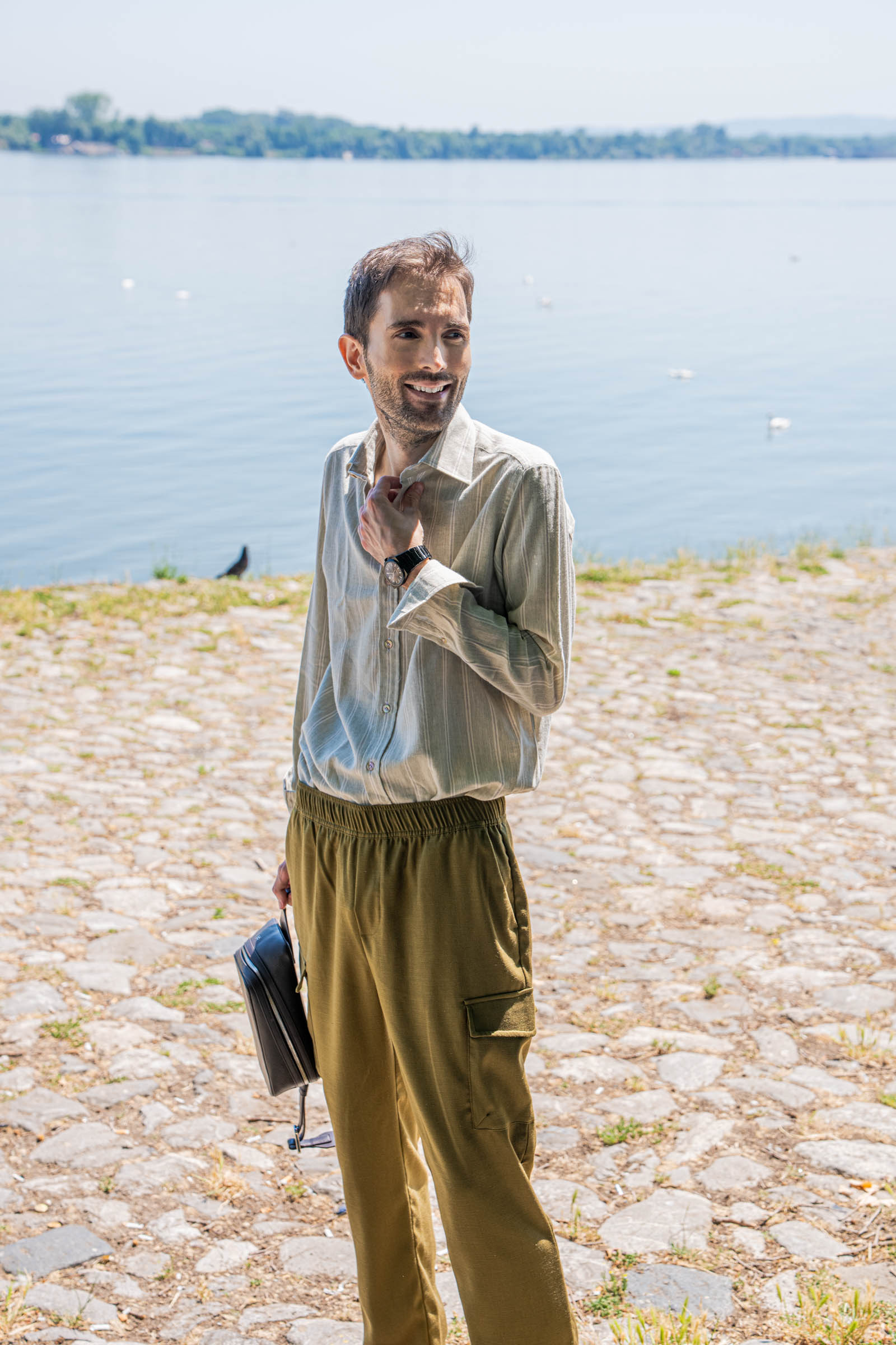 Menswear blogger Marko Tadić adjusts the collar of his Giannetto Portofino washed cotton-wool striped shirt in sage green, holding the 1900 grained leather crossbody bag beside the calm Danube river in Belgrade, Serbia.