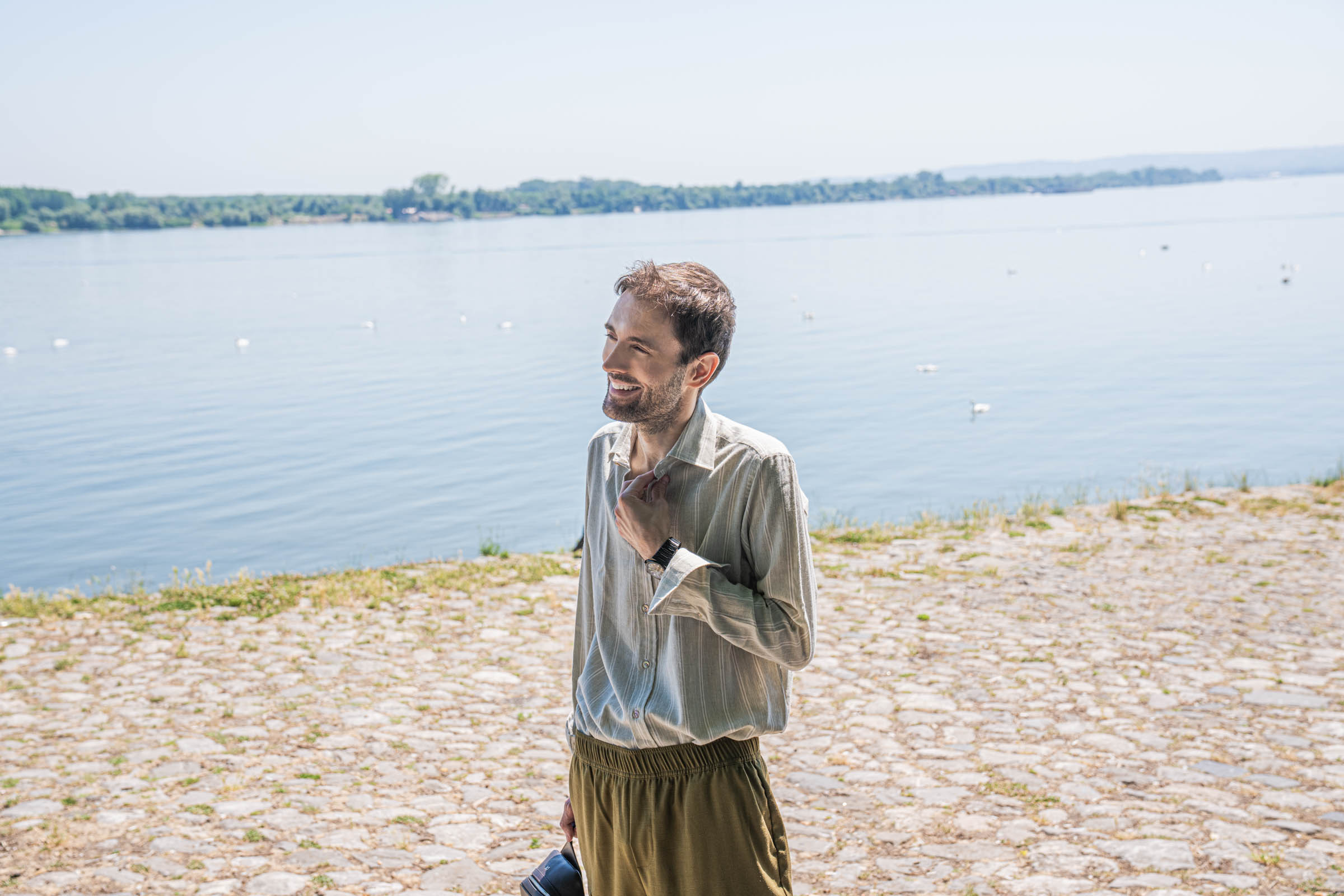 Portrait of Marko Tadić, founder of Mr.M by Marko Tadic, smiling by the Danube river in a sage green striped Giannetto Portofino shirt with a white collar, hand tucked into olive Loro Piana cargo trousers.