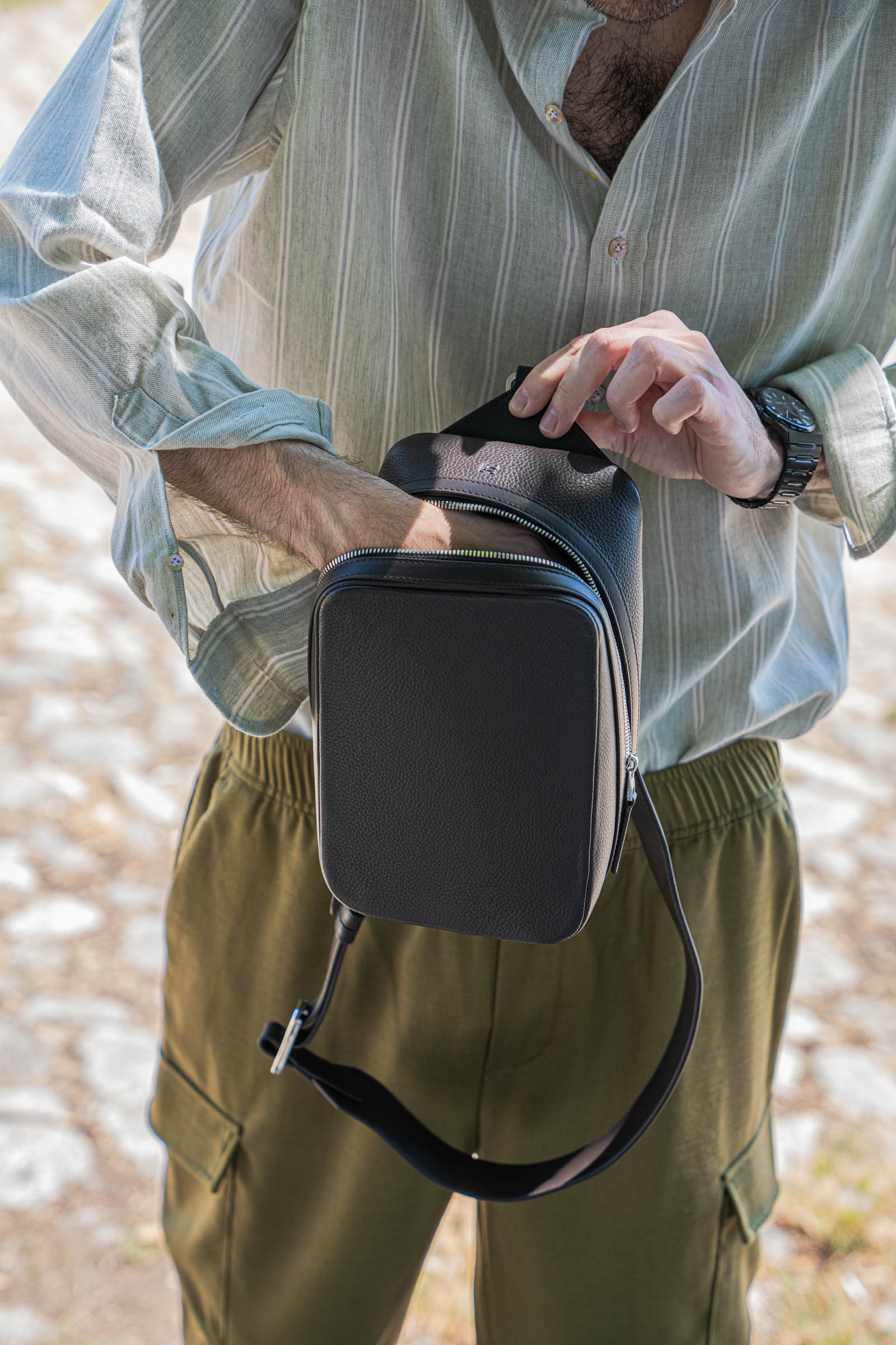 Detail shot of hands opening the main compartment of the 1900 Tempo Grande crossbody bag, revealing the signature angled zipper, metal monogram and pebbled grained leather texture of this Swiss-engineered men's bag.