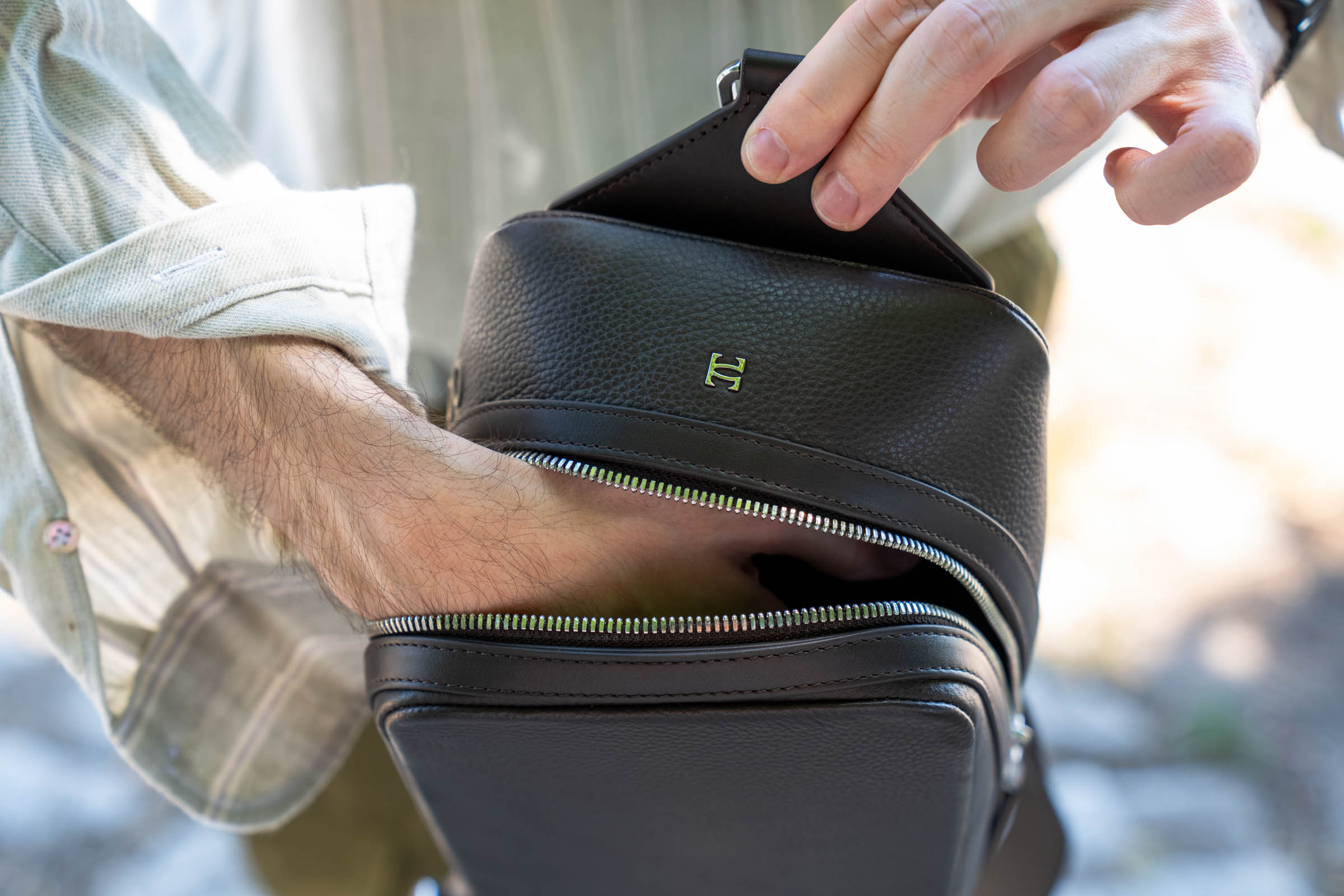 Close-up of the 1900 Tempo Grande crossbody bag showing the embossed 1900 logo on dark brown grained leather, polished silver double zippers and the brushed leather top panel, photographed on menswear blogger Marko Tadić.