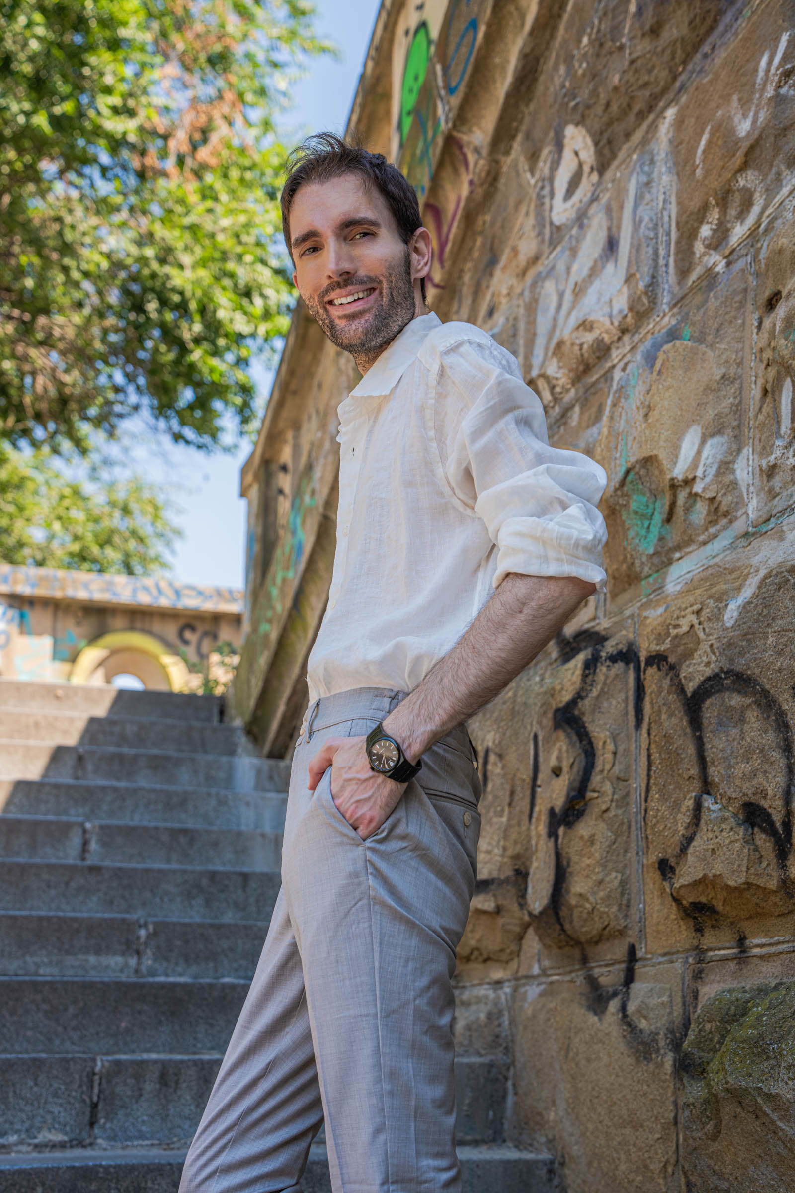 Marko Tadić, founder of the Mr.M blog, with crossed arms in a white linen Barena Venezia shirt and beige trousers against a graffiti stone wall in Belgrade
