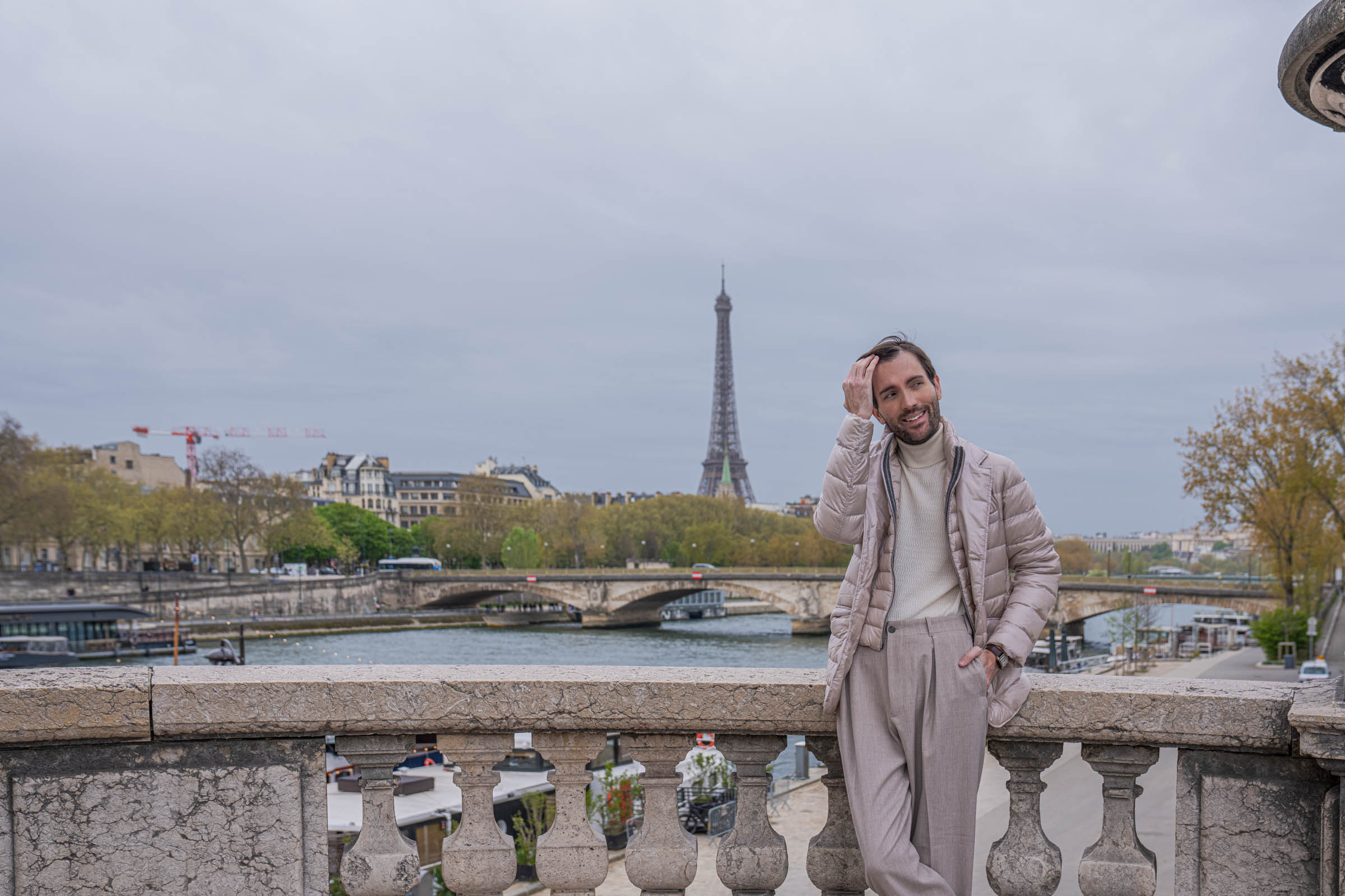 Wave-textured cream cashmere Armani turtleneck and pleated beige trousers, with a pearl beige Herno down jacket held casually over one shoulder above the Seine