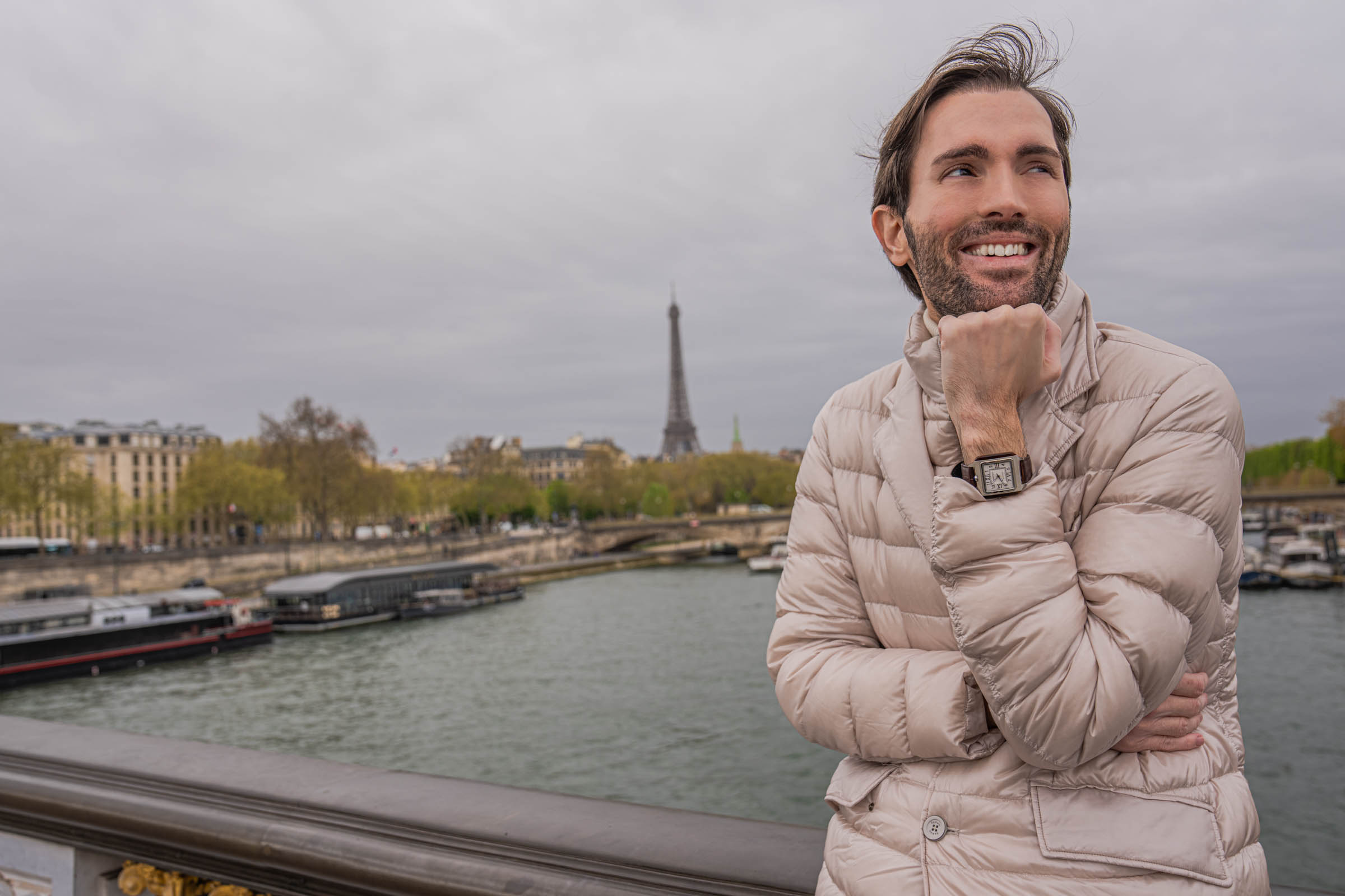 Side profile of a man smiling over his shoulder with the collar of his pearl beige Herno down jacket popped, the Eiffel Tower and spring trees lining the Seine behind