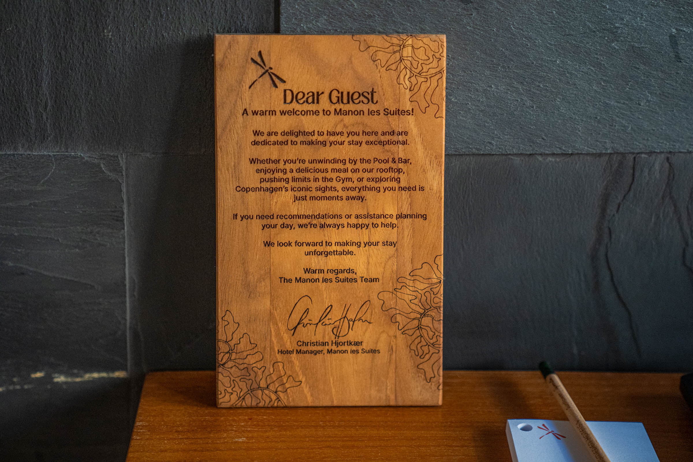 Engraved wooden welcome board from the Manon Les Suites team signed by hotel manager Christian Hjortkær on a teak desk