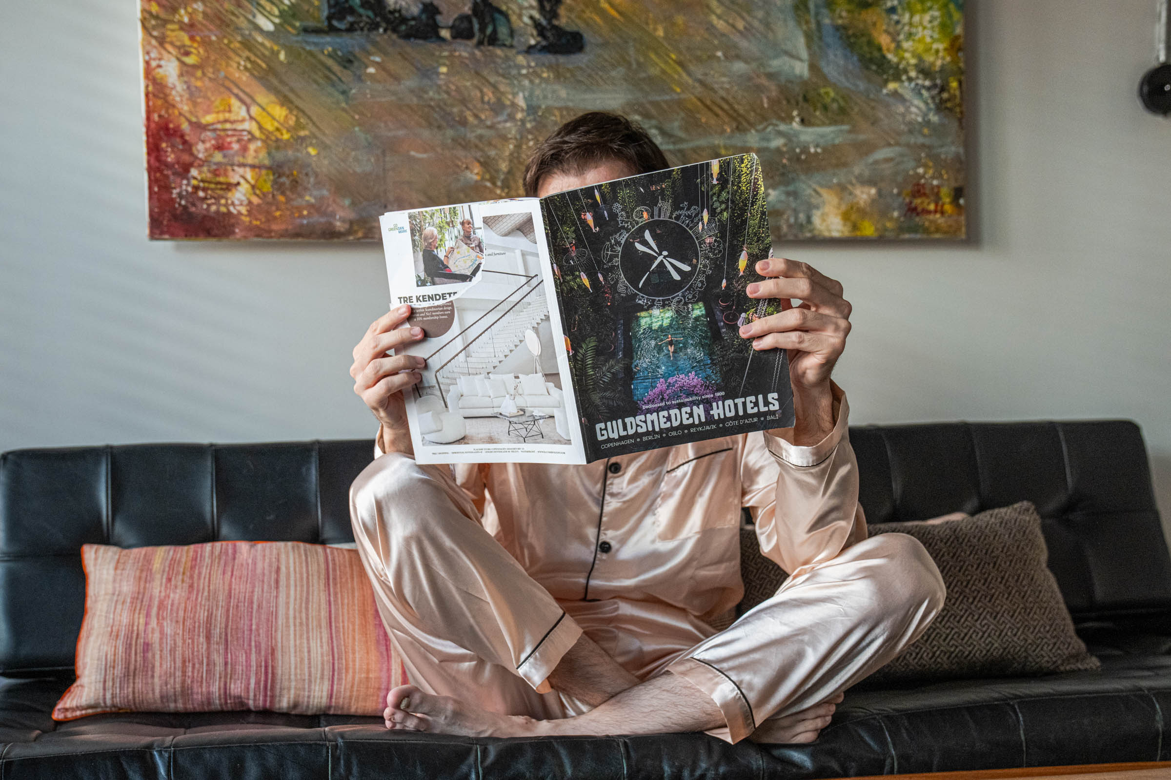 Marko Tadić in champagne silk pyjamas reading the Guldsmeden Hotels magazine on a black leather daybed in his Copenhagen suite