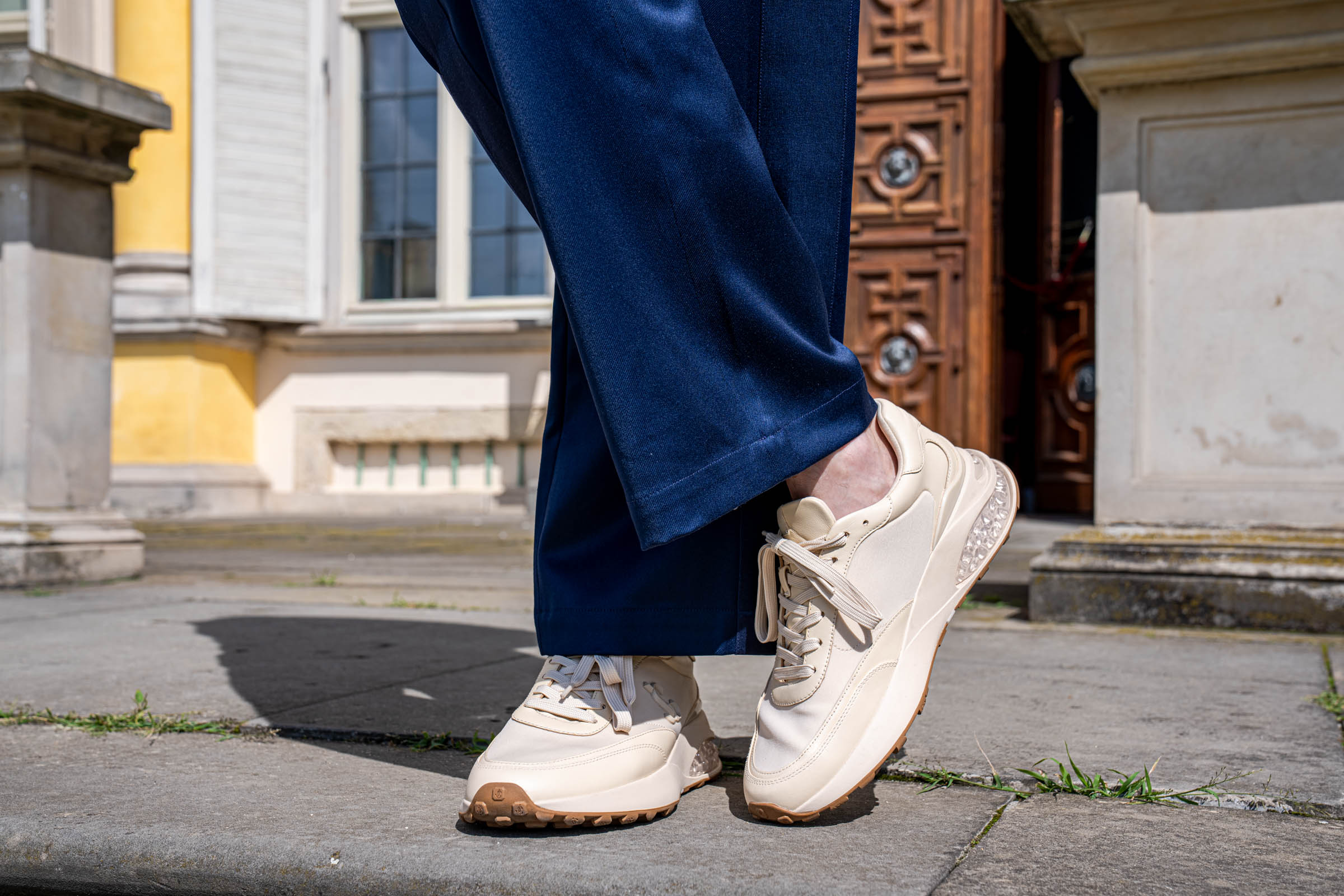 Cream Jimmy Choo Memphis sneakers with crystal-effect heel inserts and gum sole, worn under navy wide-leg trousers.