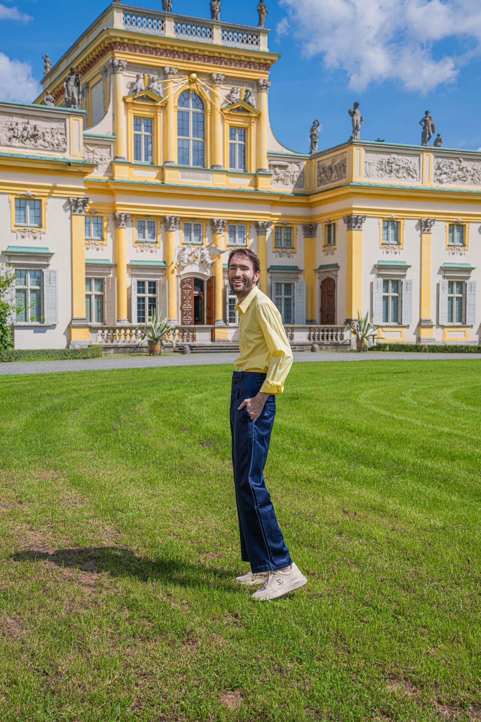 Side view of Marko Tadić on the Wilanów garden lawn, showing the wide leg and satin side piping of his navy BLUEMARBLE trousers.