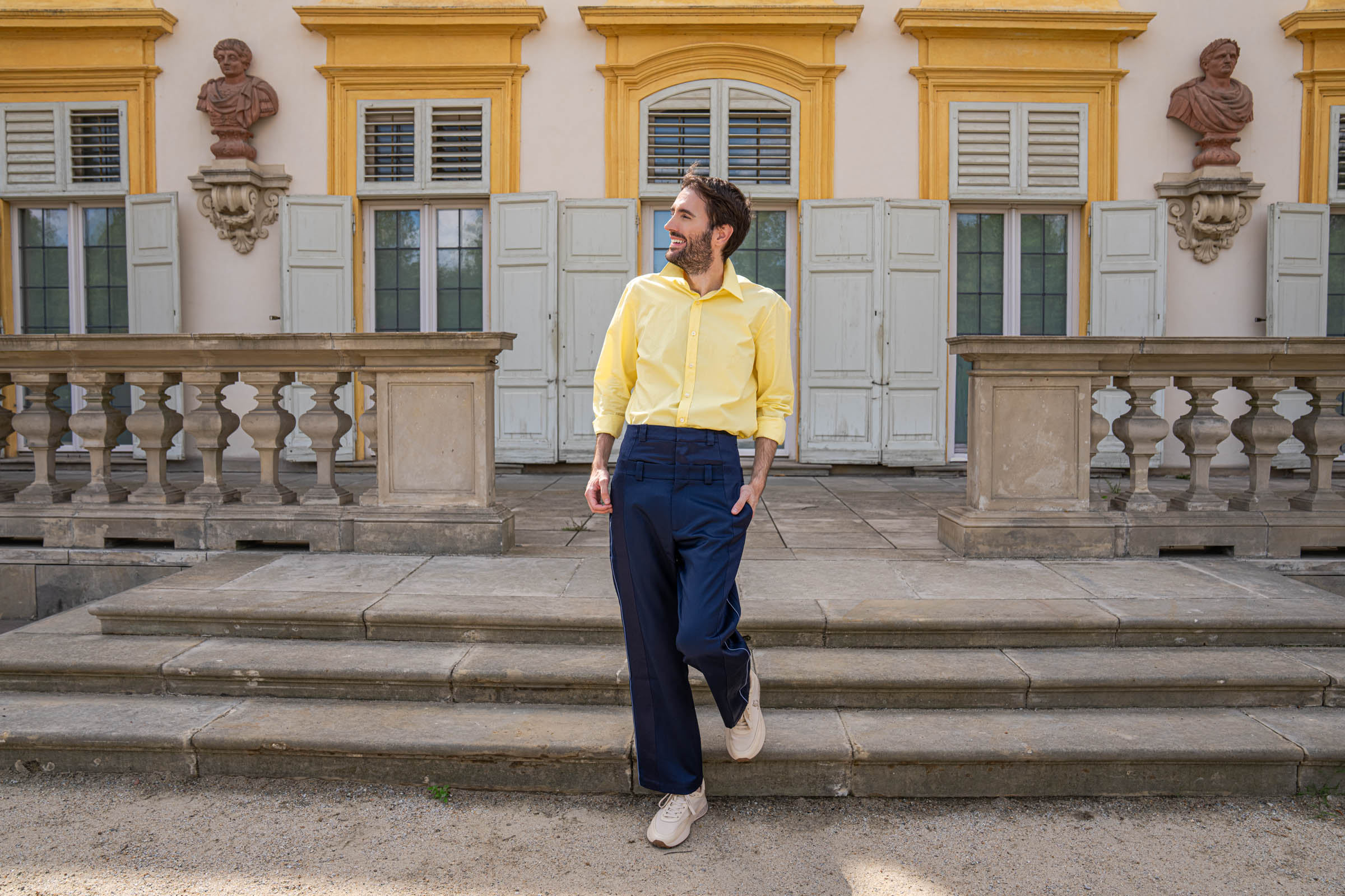 Marko Tadić poses on the stone steps of Wilanów Palace, hands in the pockets of his navy double-waist trousers.