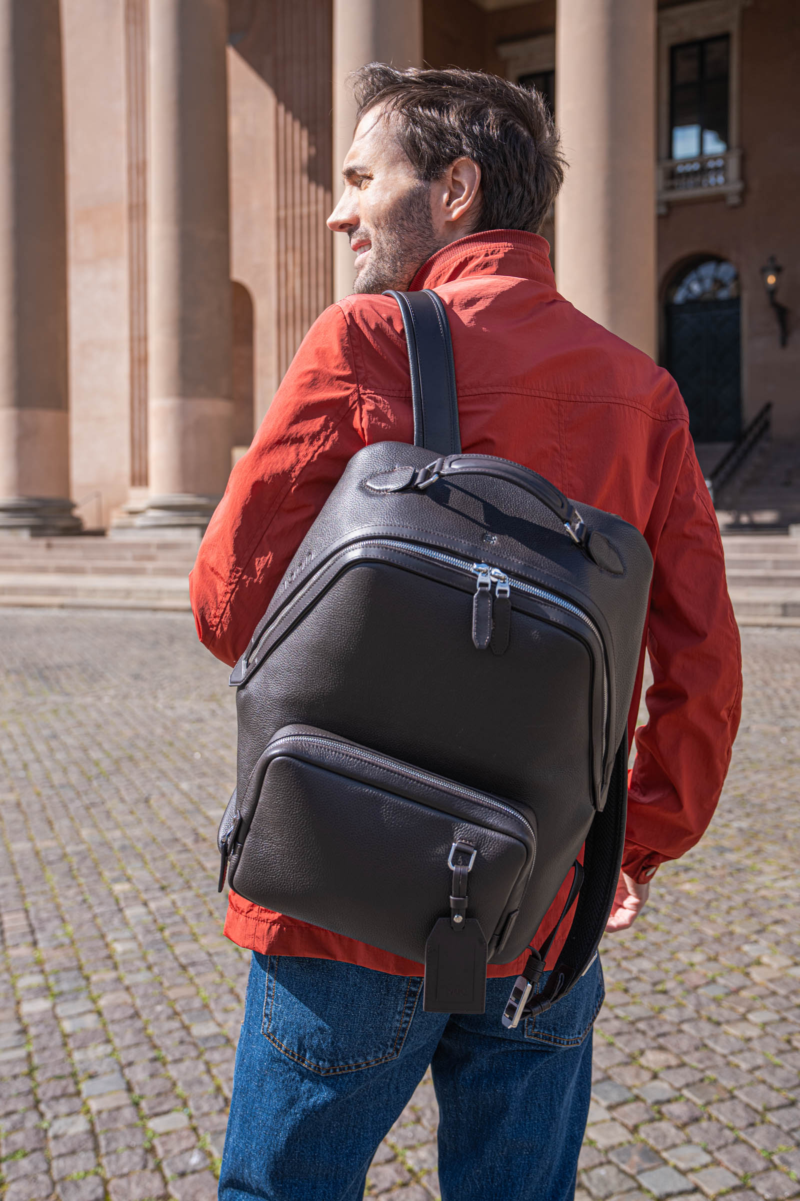 Men's travel outfit in Copenhagen: Marko Tadić in a red Canali field jacket and dark brown leather backpack beside the historic courthouse on Nytorv