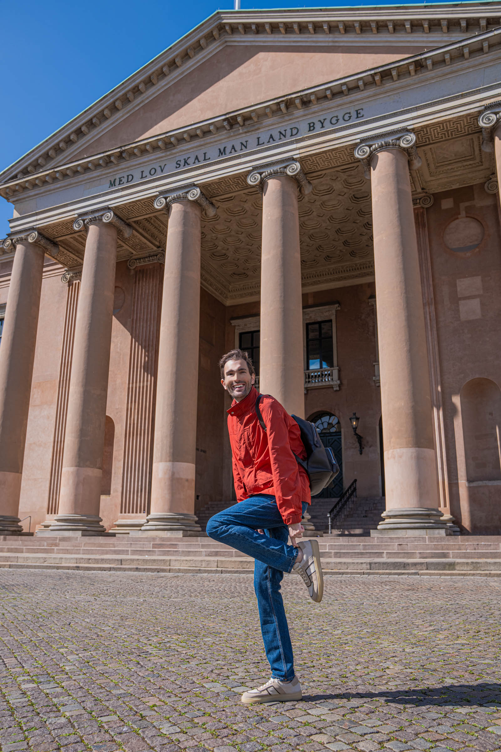 Men's smart casual look on Nytorv square, with Marko Tadić holding the 1900 Tempo Grande grained leather backpack in a red Canali field jacket and Versace denim