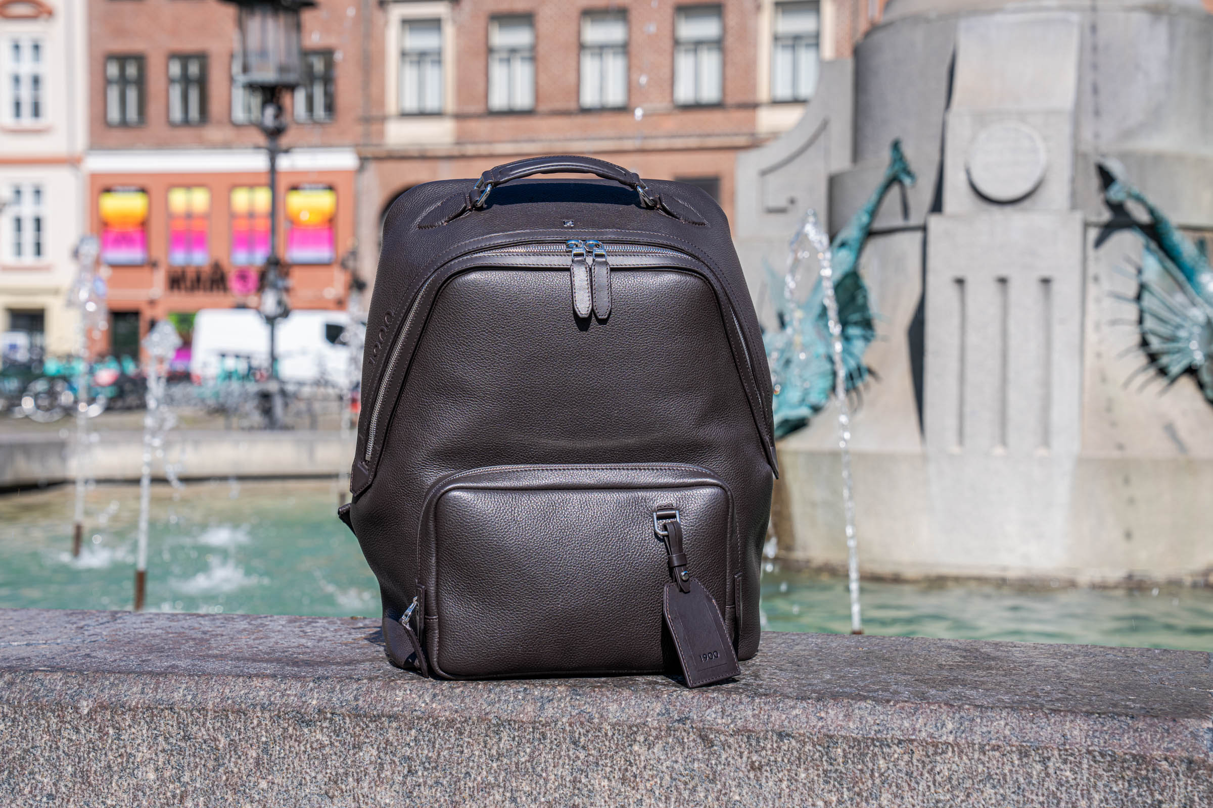 Close up of the dark brown 1900 Tempo Grande leather backpack carried by its padded top handle, showing the angled zip and removable travel tag
