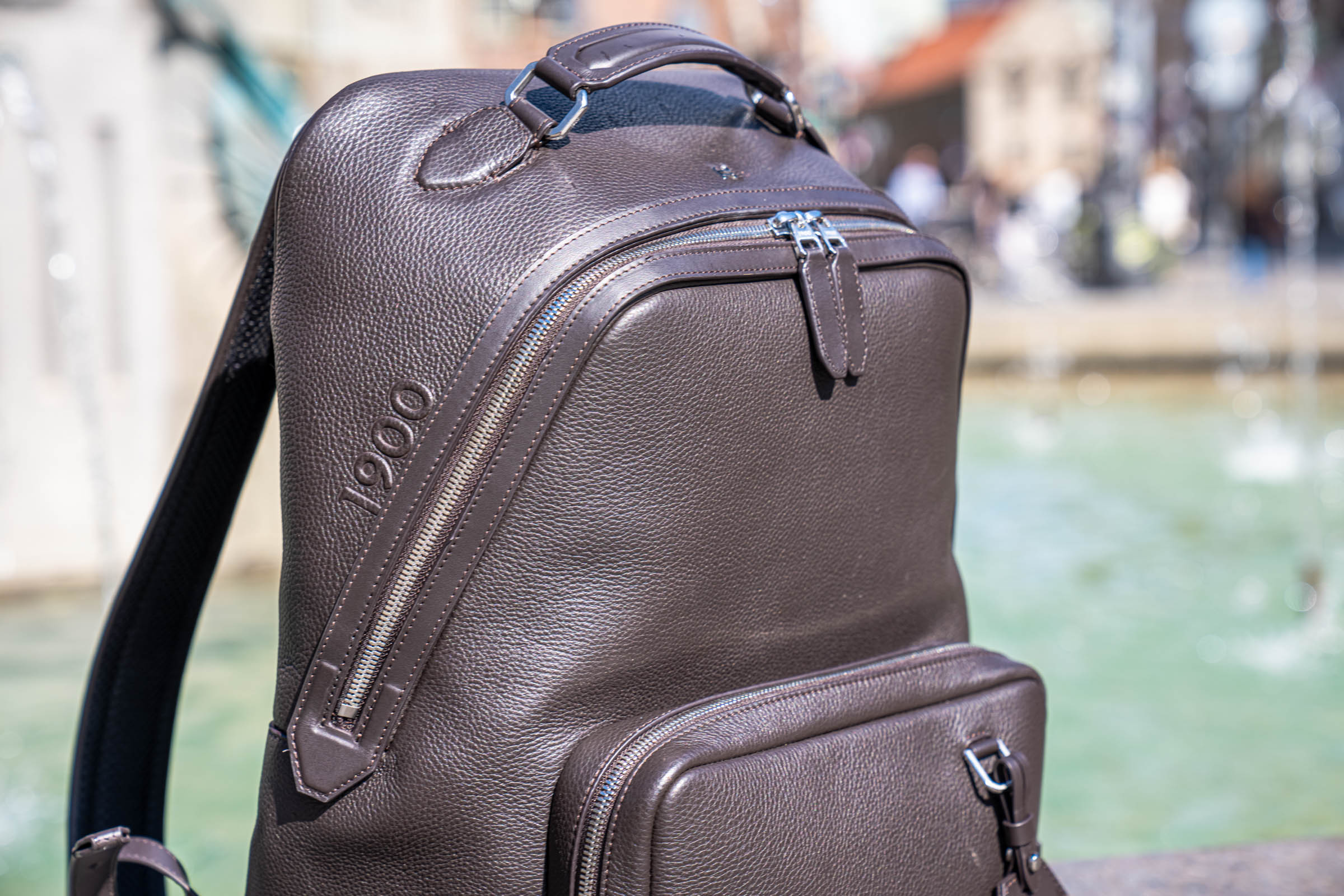 Close up of the embossed 1900 logo and signature angled zip closure on the side panel of the Tempo Grande grained leather backpack
