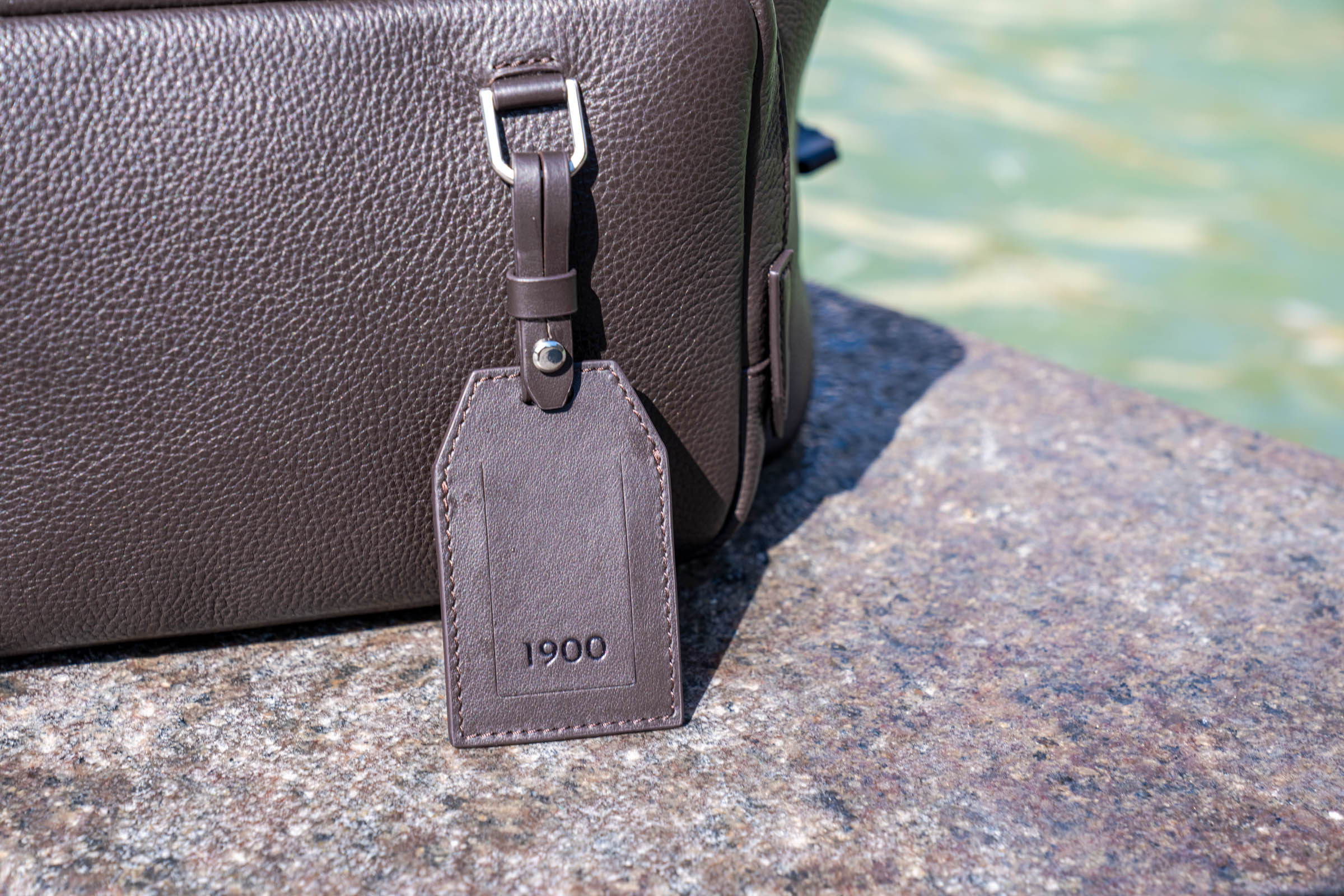 Removable leather luggage tag embossed with the 1900 logo, attached to the dark brown Tempo Grande backpack on granite stone in Copenhagen