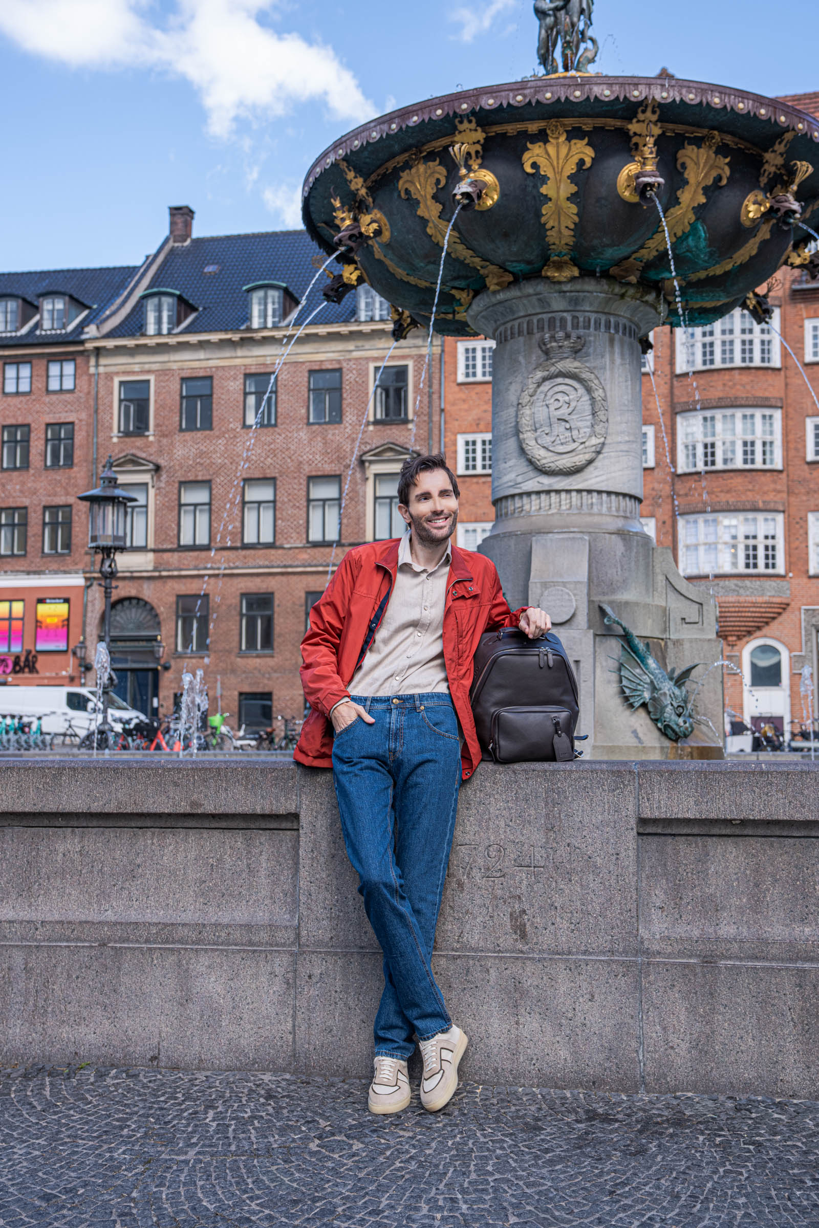 Marko Tadić, founder of Mr.M blog, wearing a red Canali field jacket and blue Versace jeans while leaning on the Caritas Fountain at Gammeltorv square in Copenhagen