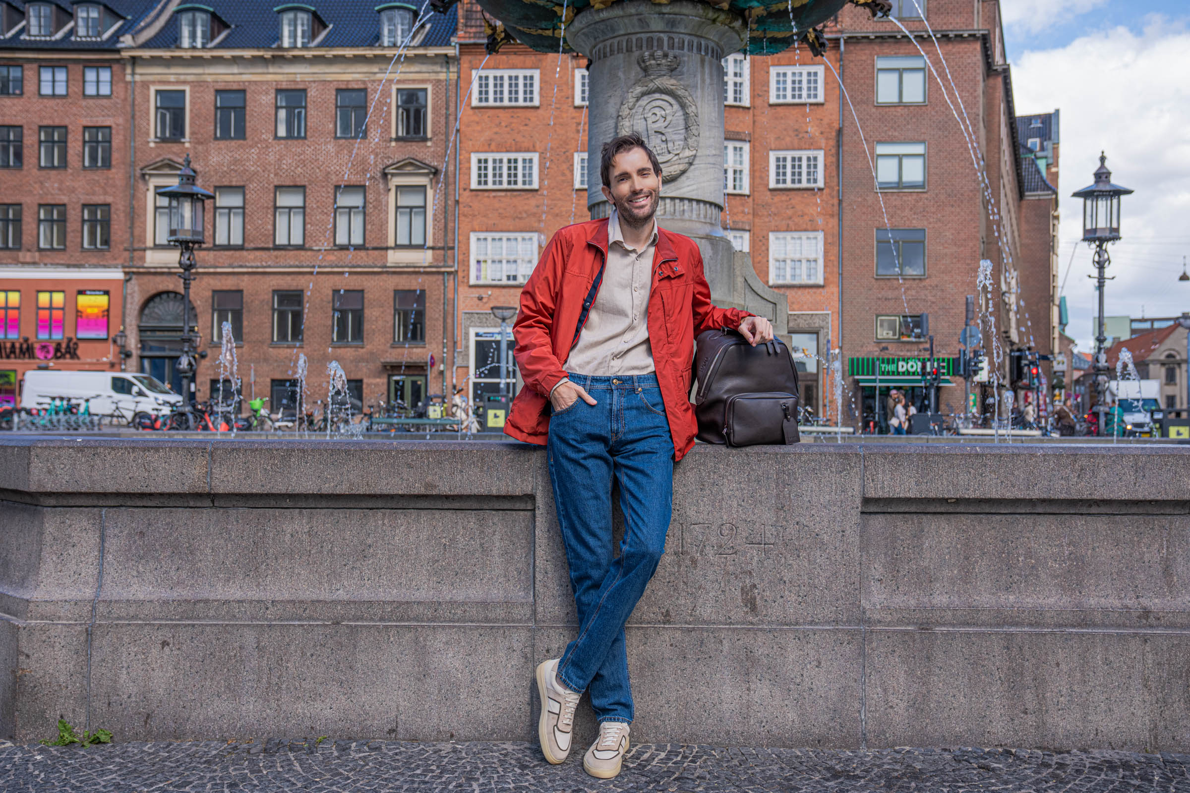 Luxury menswear editorial in Copenhagen showing Marko Tadić in an open red Canali field jacket, beige washed flannel shirt and straight leg jeans near the Caritas Fountain