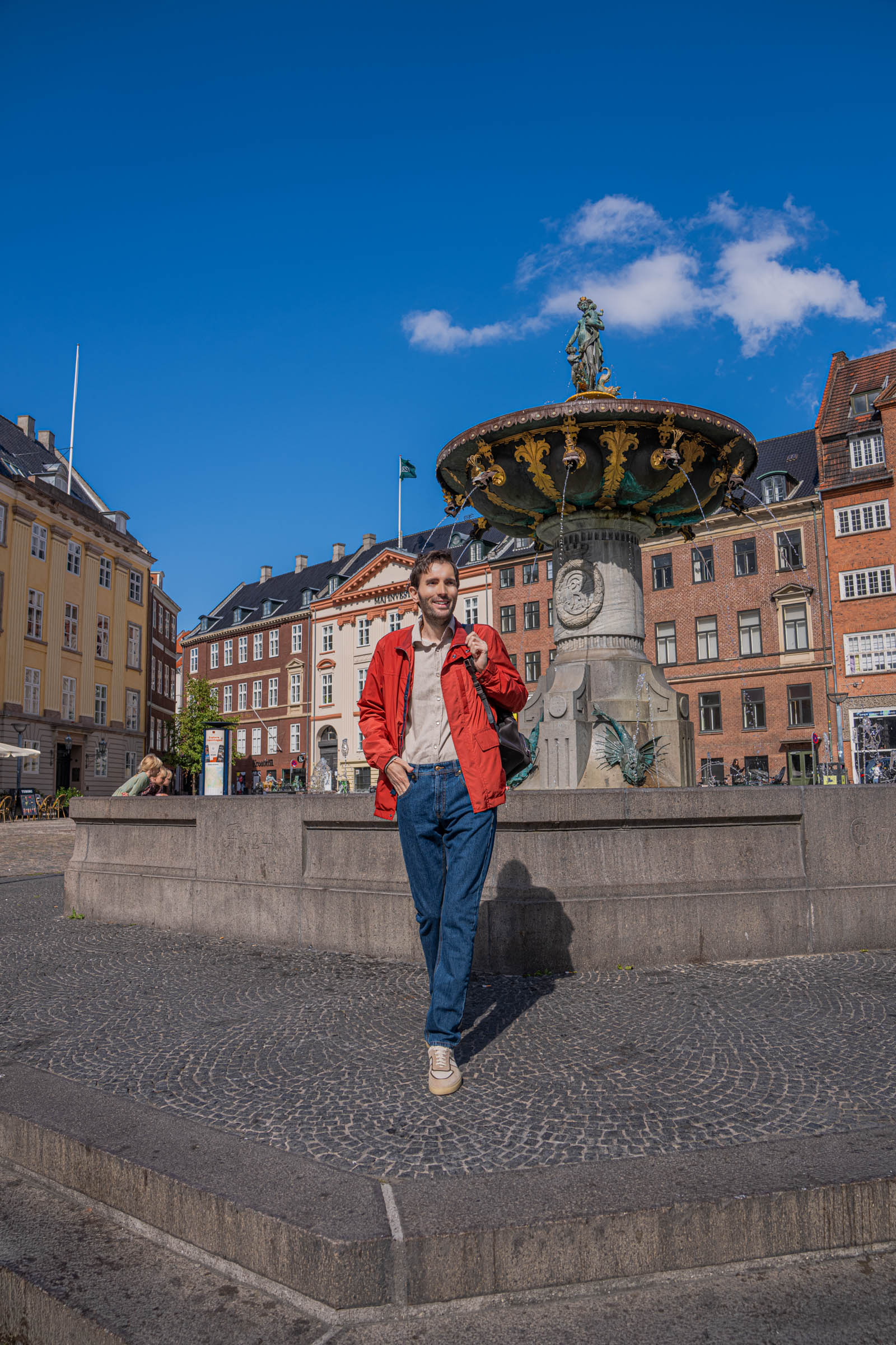 Marko Tadić standing below the bronze Caritas figure group of Copenhagen's oldest fountain, dressed in a red Canali field jacket, straight Versace denim and cream suede sneakers