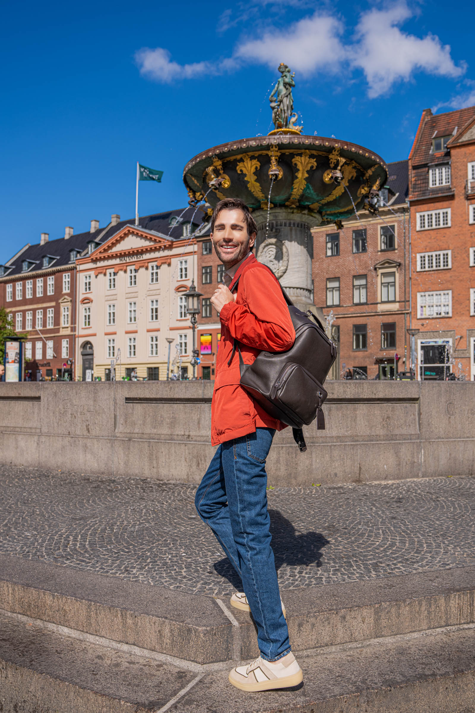 Side view of Marko Tadić carrying the 1900 Tempo Grande grained leather backpack on one shoulder, styled with a red Canali field jacket in front of the golden apple fountain