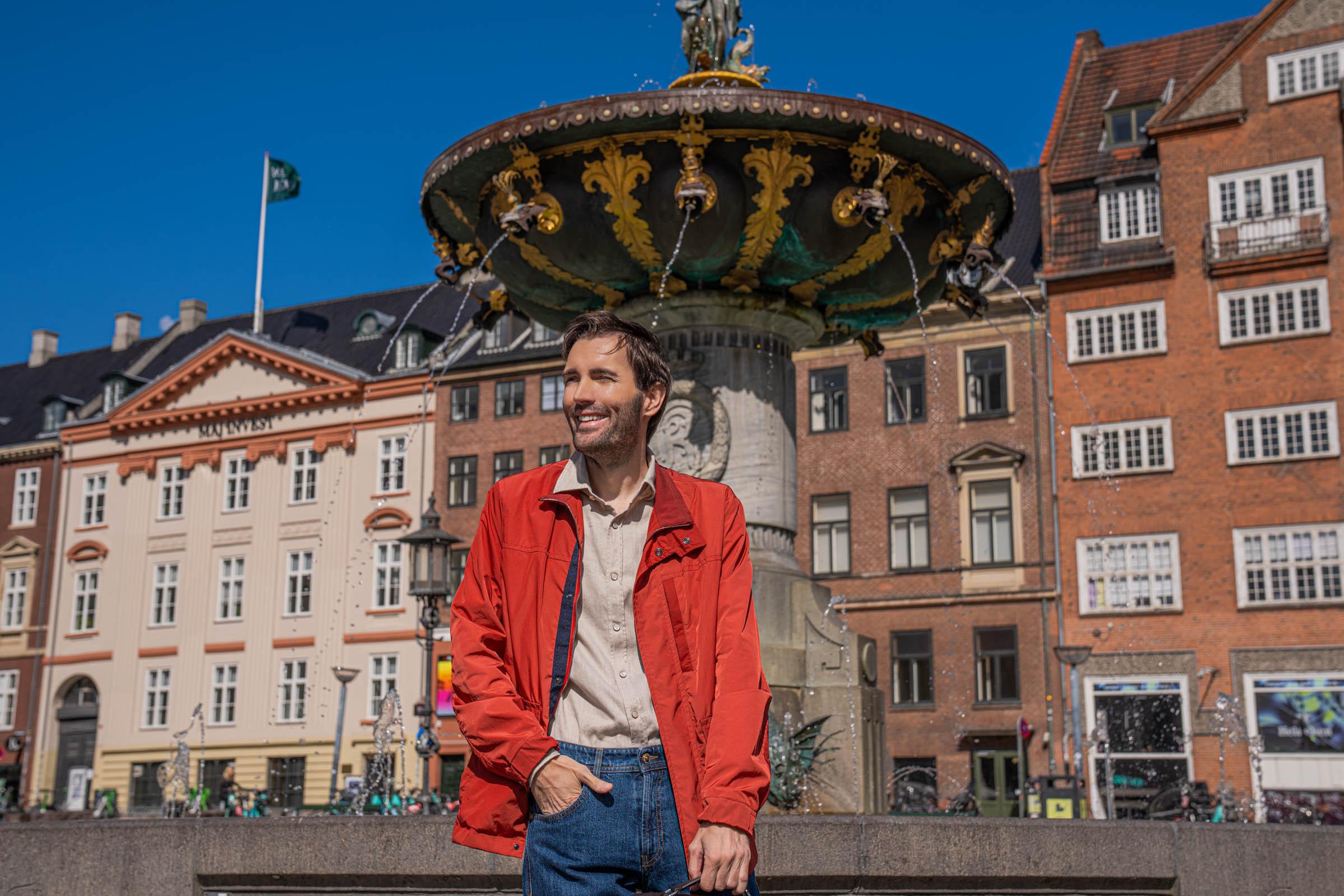 Portrait of Mr.M blog founder Marko Tadić smiling in a red Canali field jacket and beige Giannetto Portofino shirt, with Copenhagen's Renaissance fountain behind him