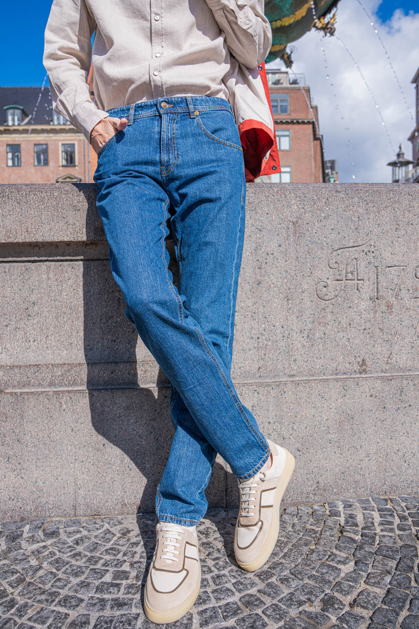 Straight leg Versace jeans in mid blue paired with cream Carlos Santos Lonnie suede sneakers, photographed against the carved granite base of Copenhagen's oldest fountain