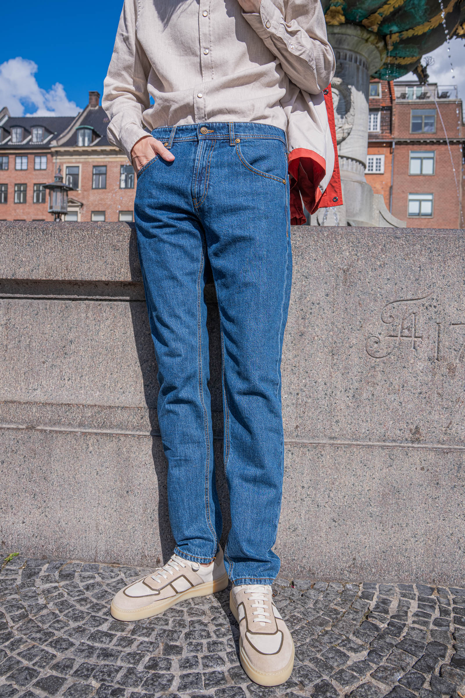 Mid blue Versace jeans with golden topstitching and a beige flannel shirt, worn by Marko Tadić beside the 1724 date carved into the Caritas Fountain in Copenhagen