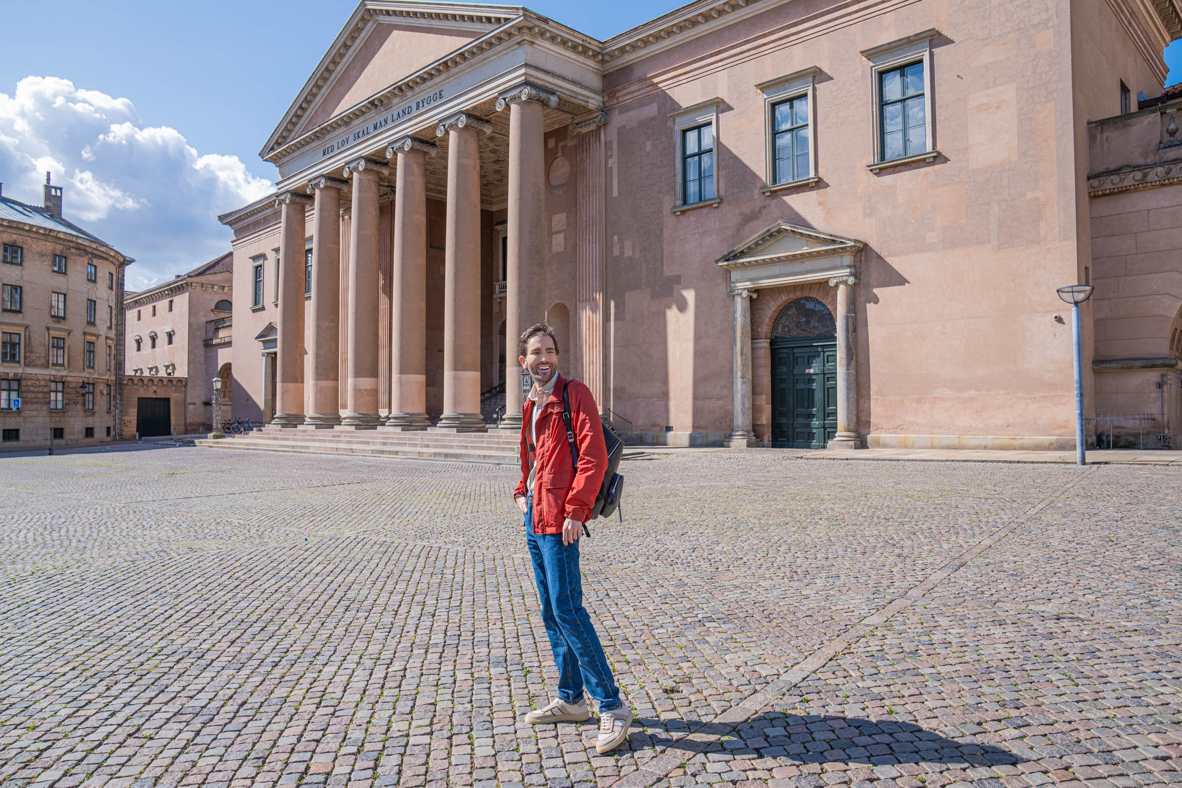Marko Tadić walking across the cobblestones of Gammeltorv square in Copenhagen under a deep blue sky, wearing a red Canali field jacket and mid blue Versace denim