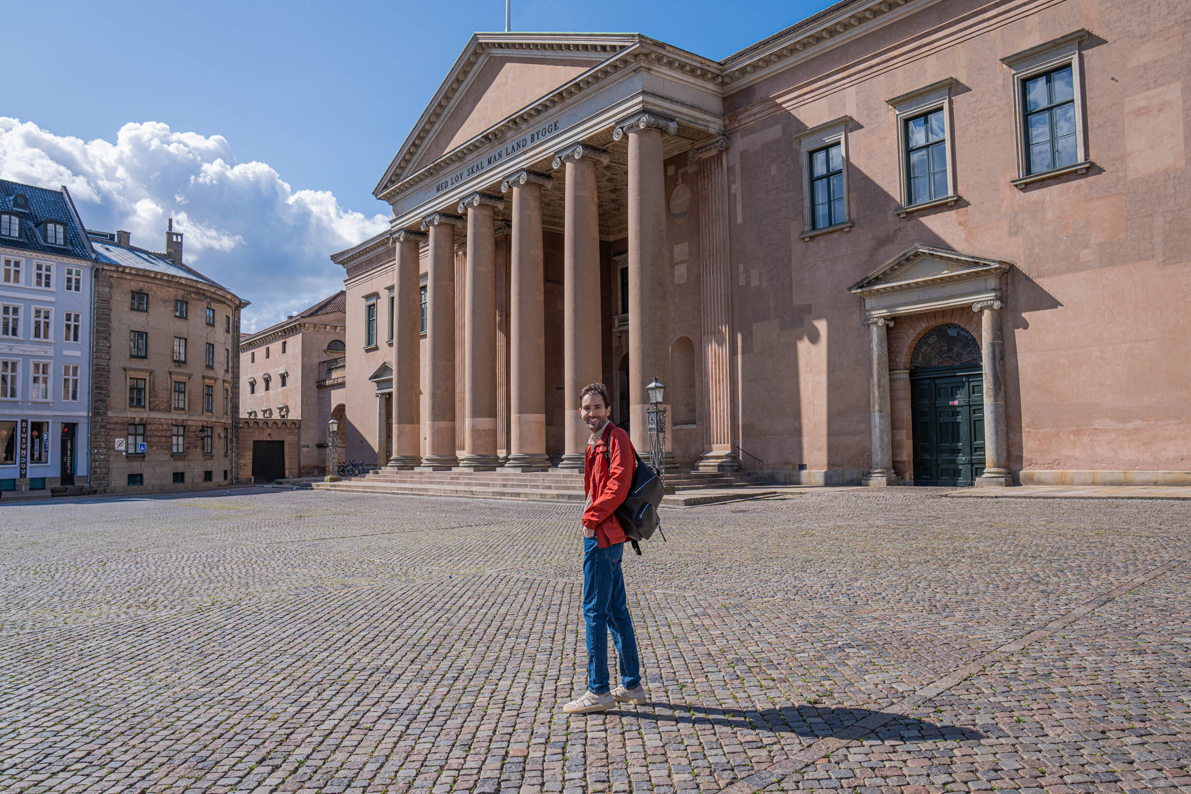 Marko Tadić walking across Nytorv square in front of the neoclassical Copenhagen Court House, wearing a red Canali field jacket and blue Versace jeans