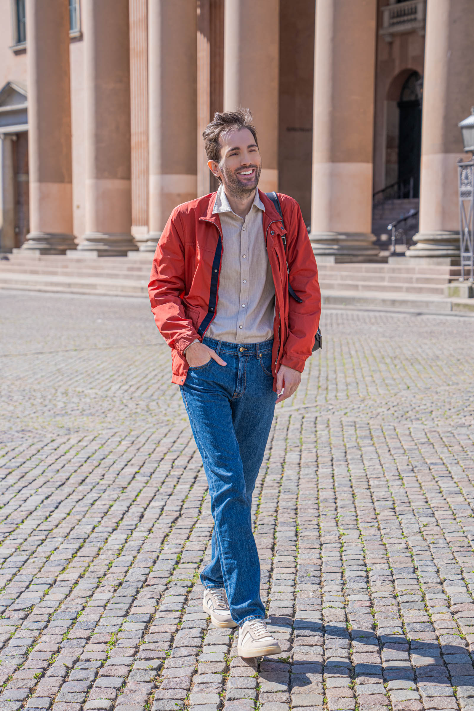 Marko Tadić walking toward the camera between the sandstone columns of Copenhagen's courthouse in a red Canali field jacket, beige flannel shirt and straight Versace jeans